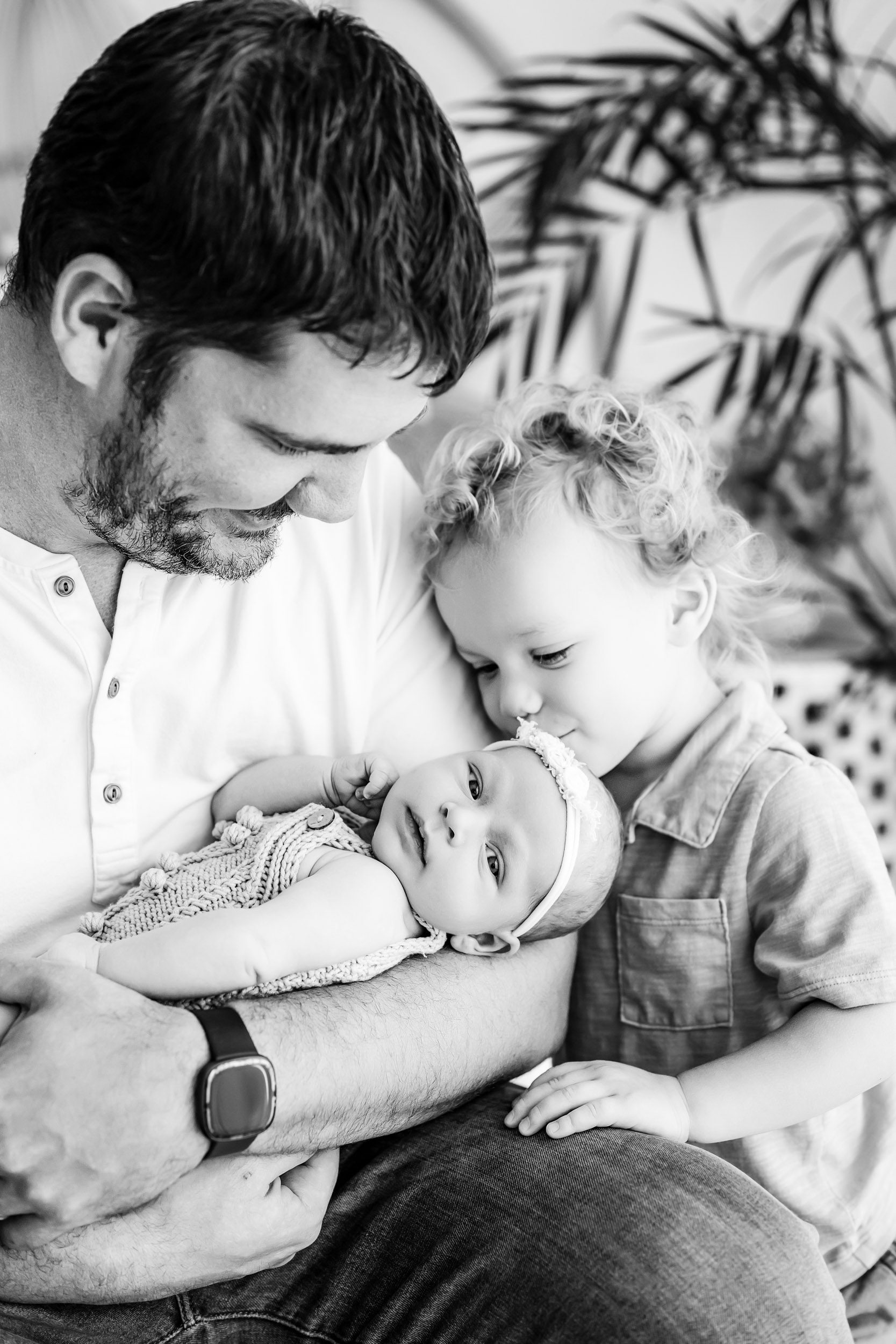 a black and white photo of a baby girl cradled in her dad's arms while her big brother gives her a kiss on the head during a newborn family photoshoot