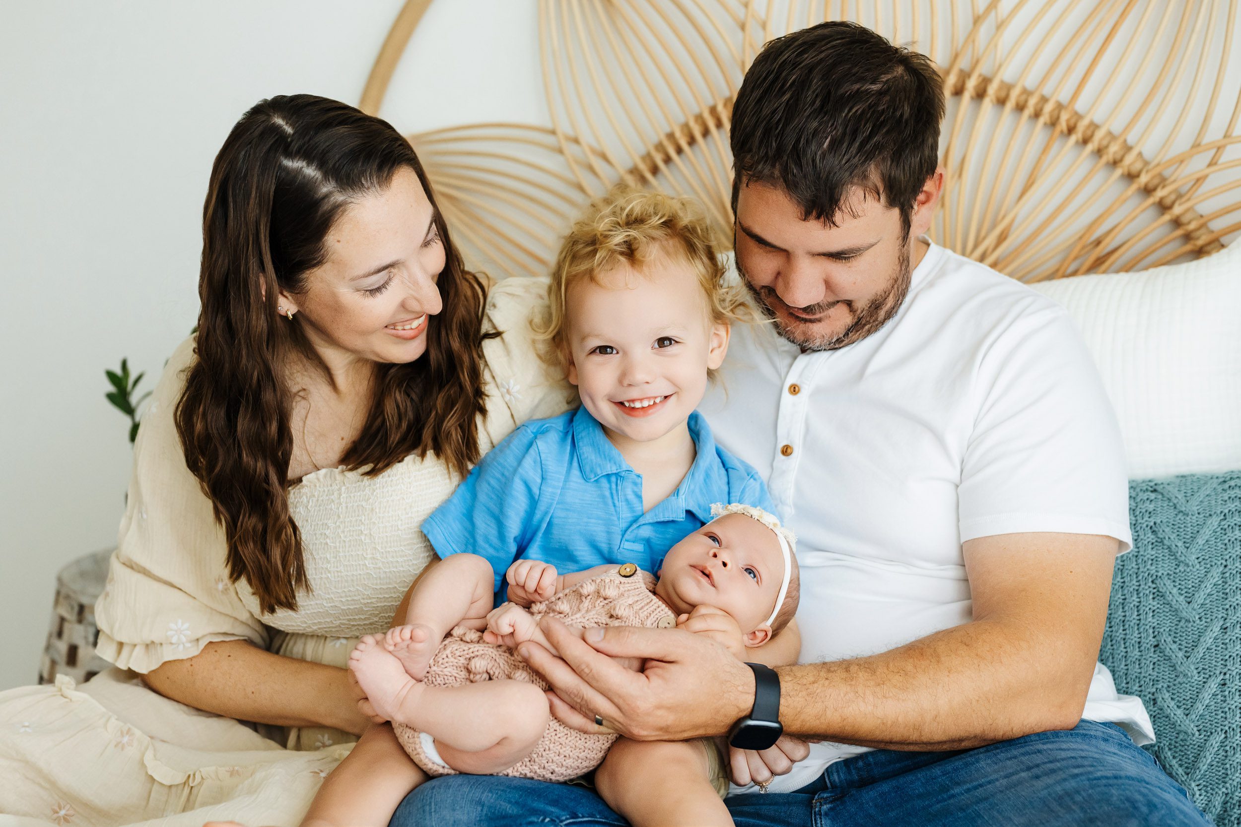 a young boy sitting on his parents lap and holding his baby sister in his arms as he looks directly at the camera with a huge smile on his face during a newborn family photoshoot