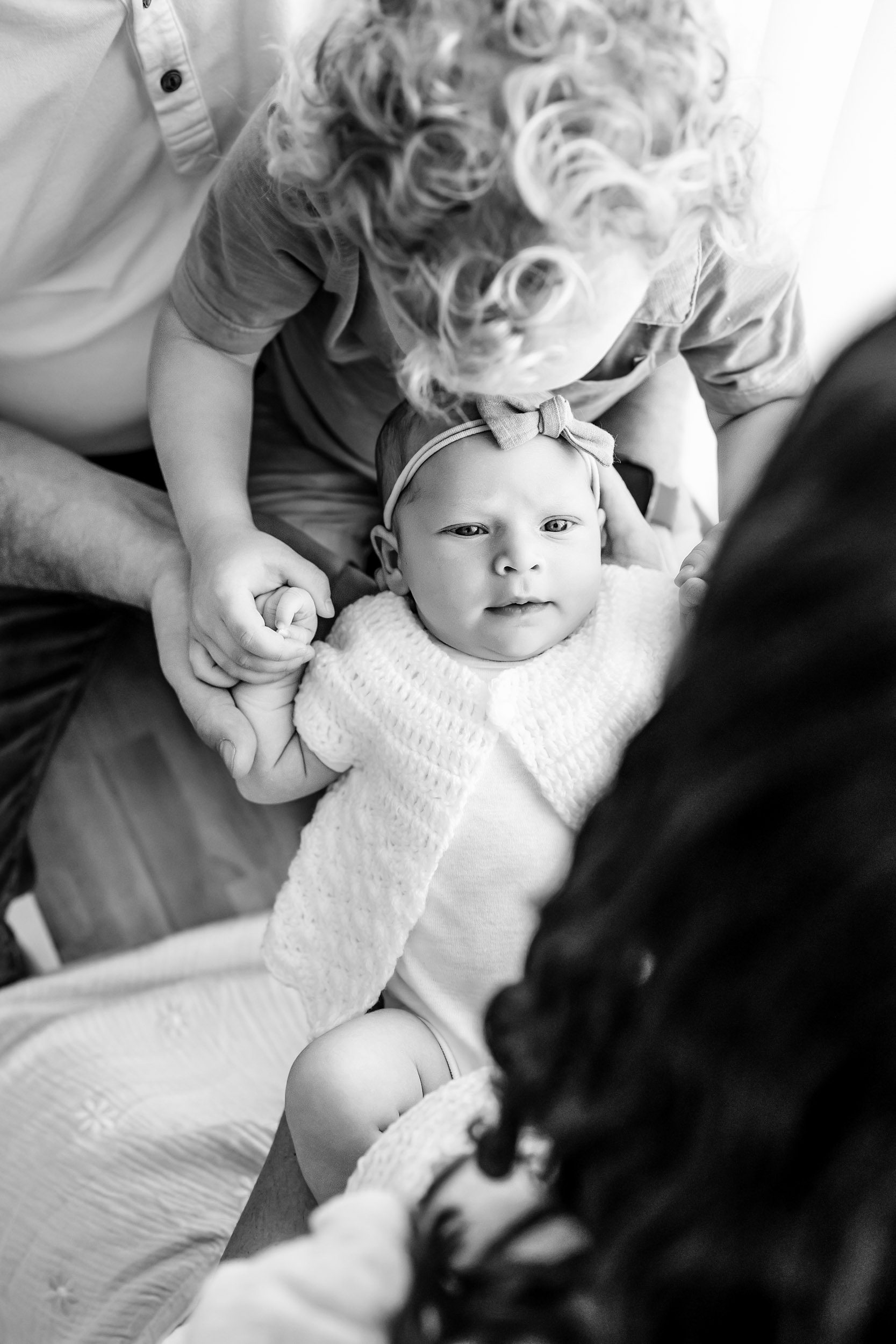 a black and white picture taken from above of a baby girl in her mom's arms as big brother gives her a kiss on the head during a newborn family photoshoot