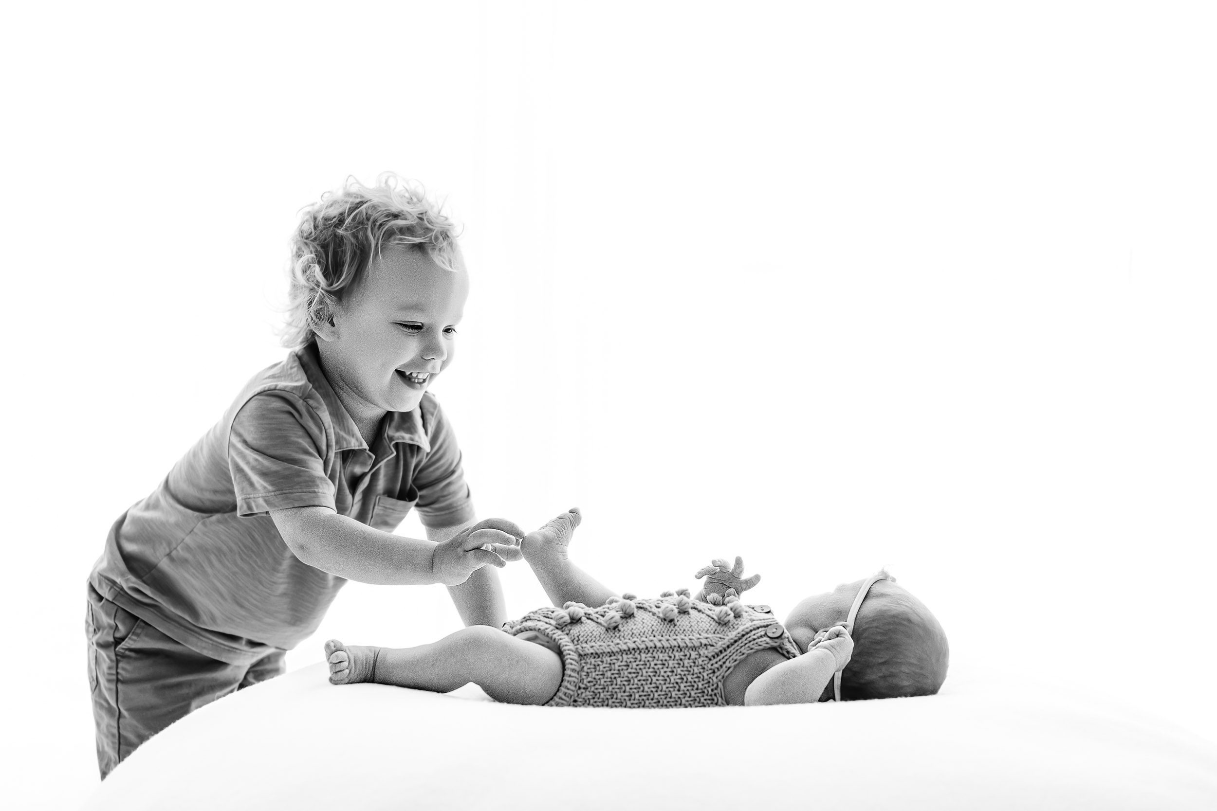 a black and white backlit photo of a baby girl laying on a bean bag as her big brother tickles her toes during a newborn family photoshoot