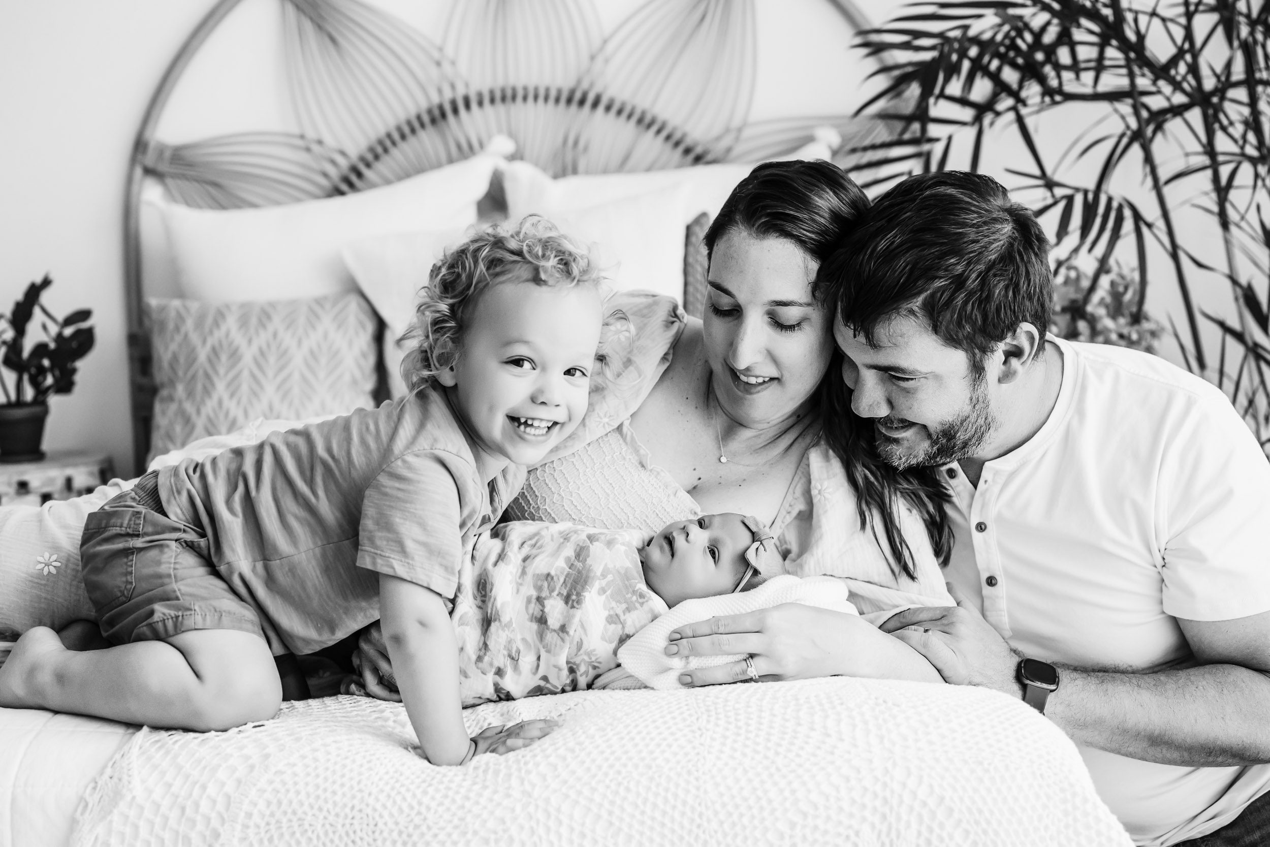a black and white picture of a family of four laying on a bed snuggling with their baby girl as their young son smiles at the camera during a newborn family photoshoot