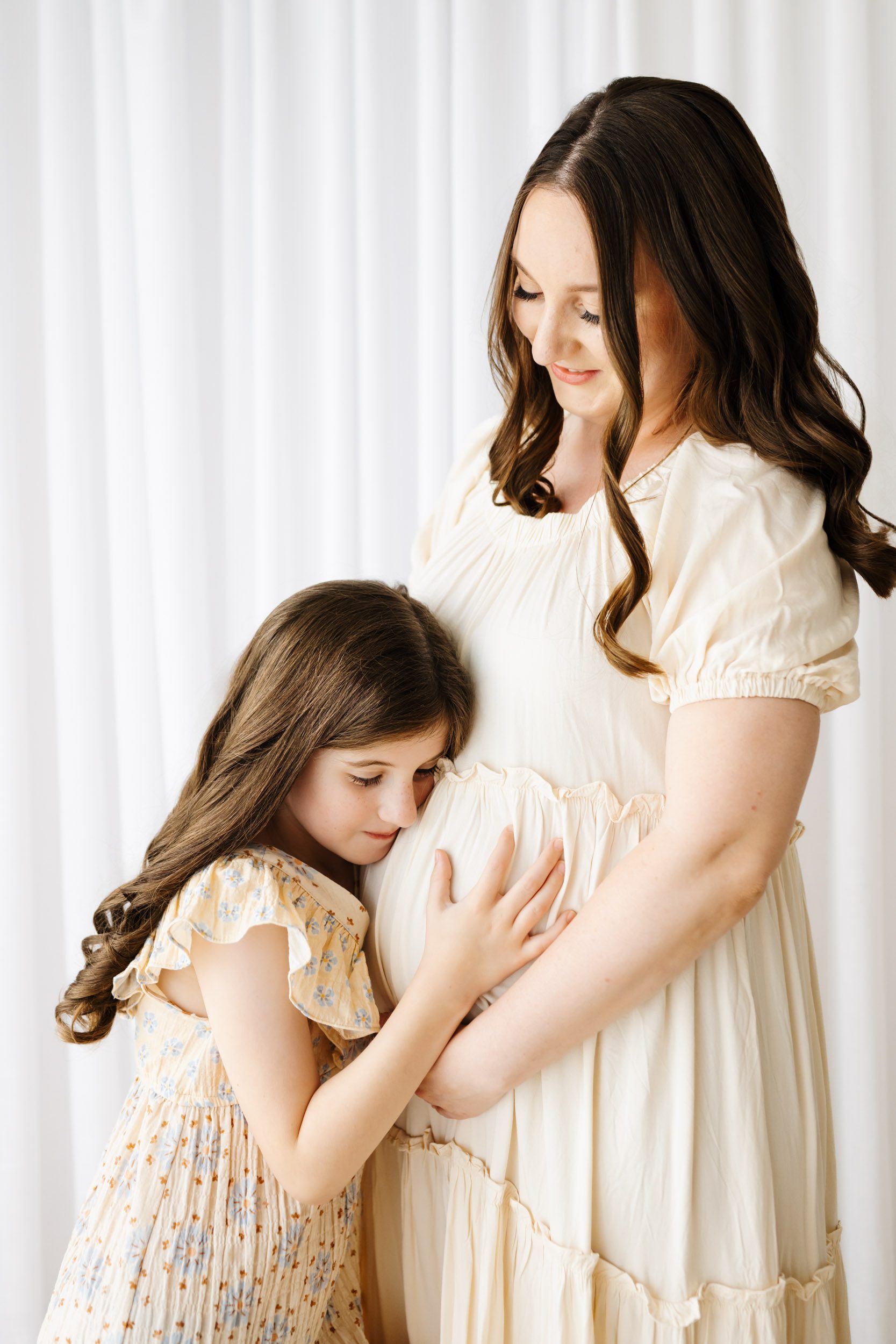 a big sister to be resting her head on her mom's belly as mom smiles down at her during a lifestyle studio maternity photoshoot