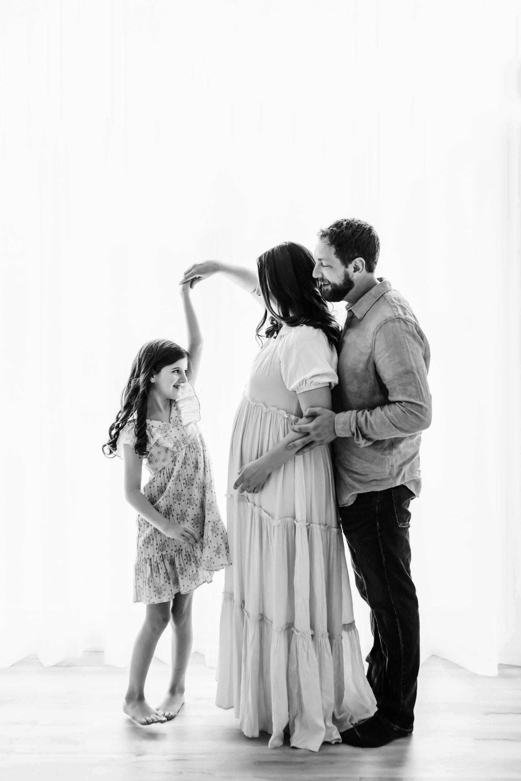 a black and white backlit photo of an expecting mom holding her daughter's hand and twirling her as she spins while dad hugs mom from behind during a lifestyle studio maternity photoshoot