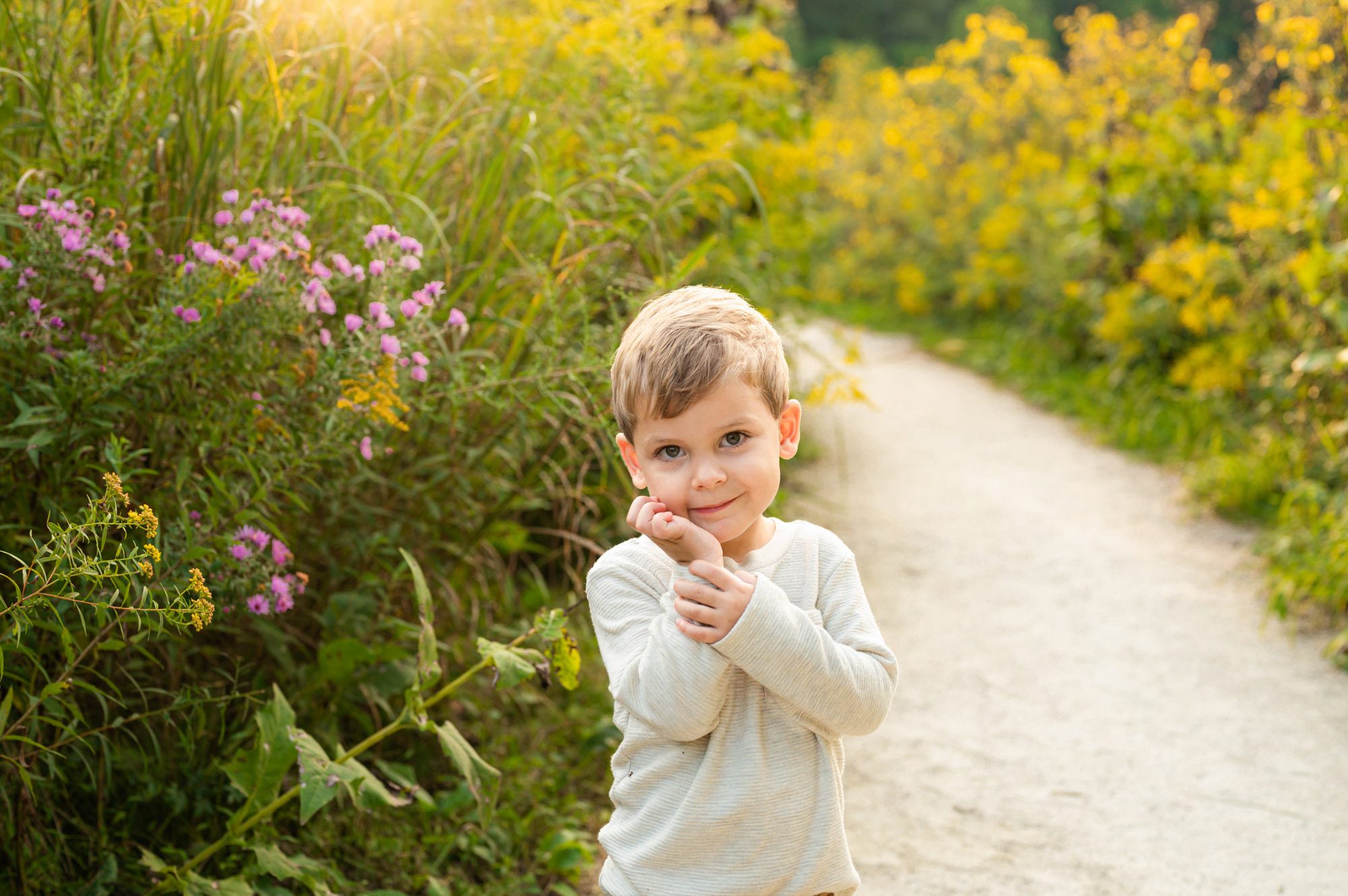 A NAPCP master baby photographer portfolio image of a young a young boy standing on a path surrounded by fields of wildflowers in bloom as he holds his hands up to his cheeks and smiles directly at the camera