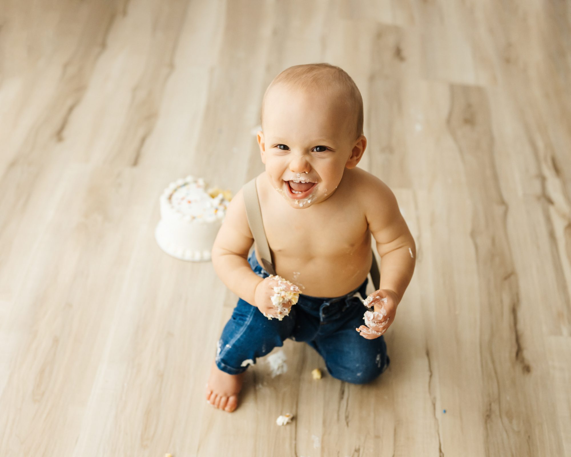 A NAPCP master baby photographer portfolio image of a young boy sitting on the floor next to a white birthday cake with icing on his face and hands as he looks up at the camera with a huge smile on his face during a 1st birthday cake smash photoshoot