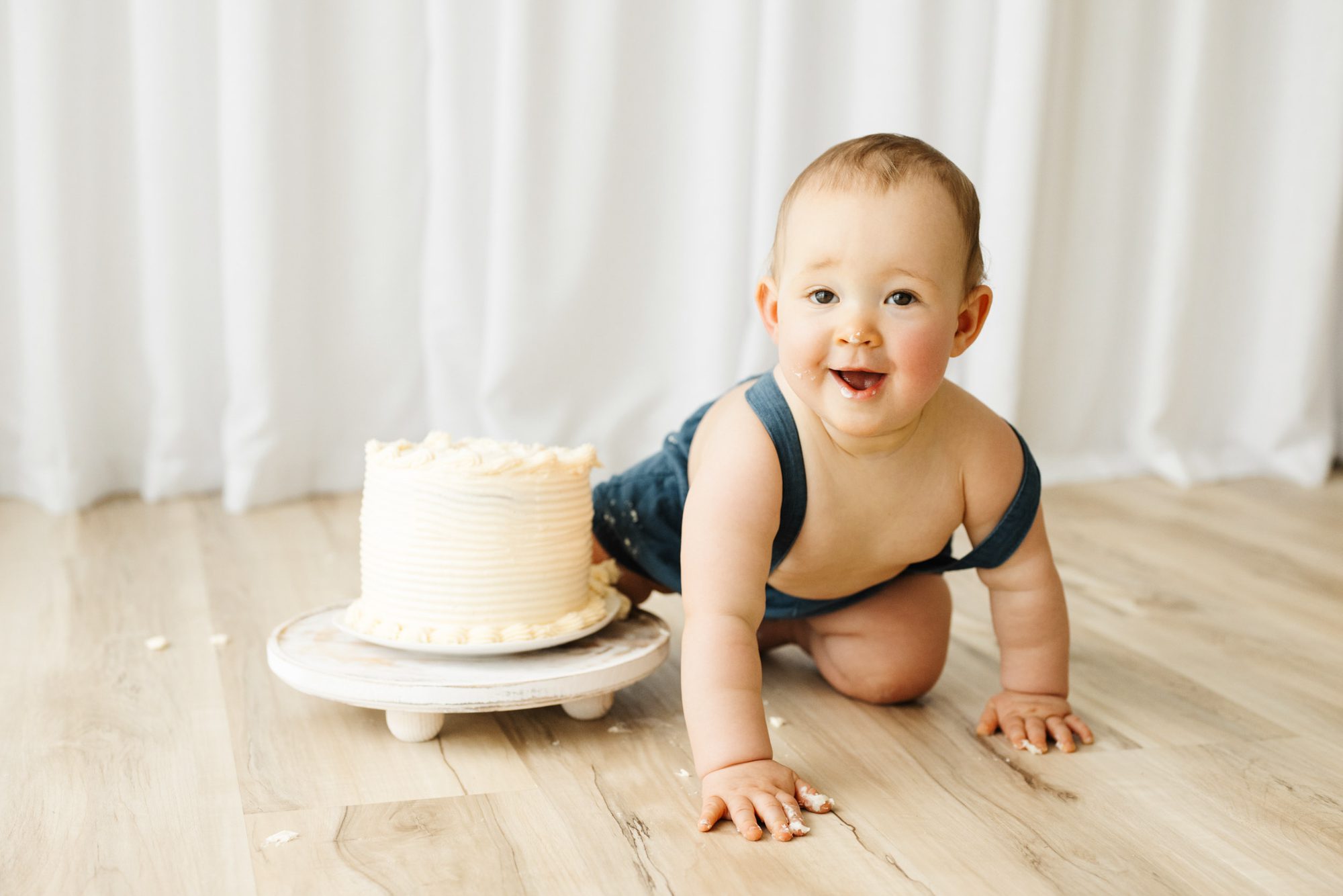 A NAPCP master baby photographer portfolio image of a young boy sitting on the floor next to a white birthday cake with icing on his face and hands as he leans forward and smiles directly at the camera during a 1st birthday cake smash photoshoot