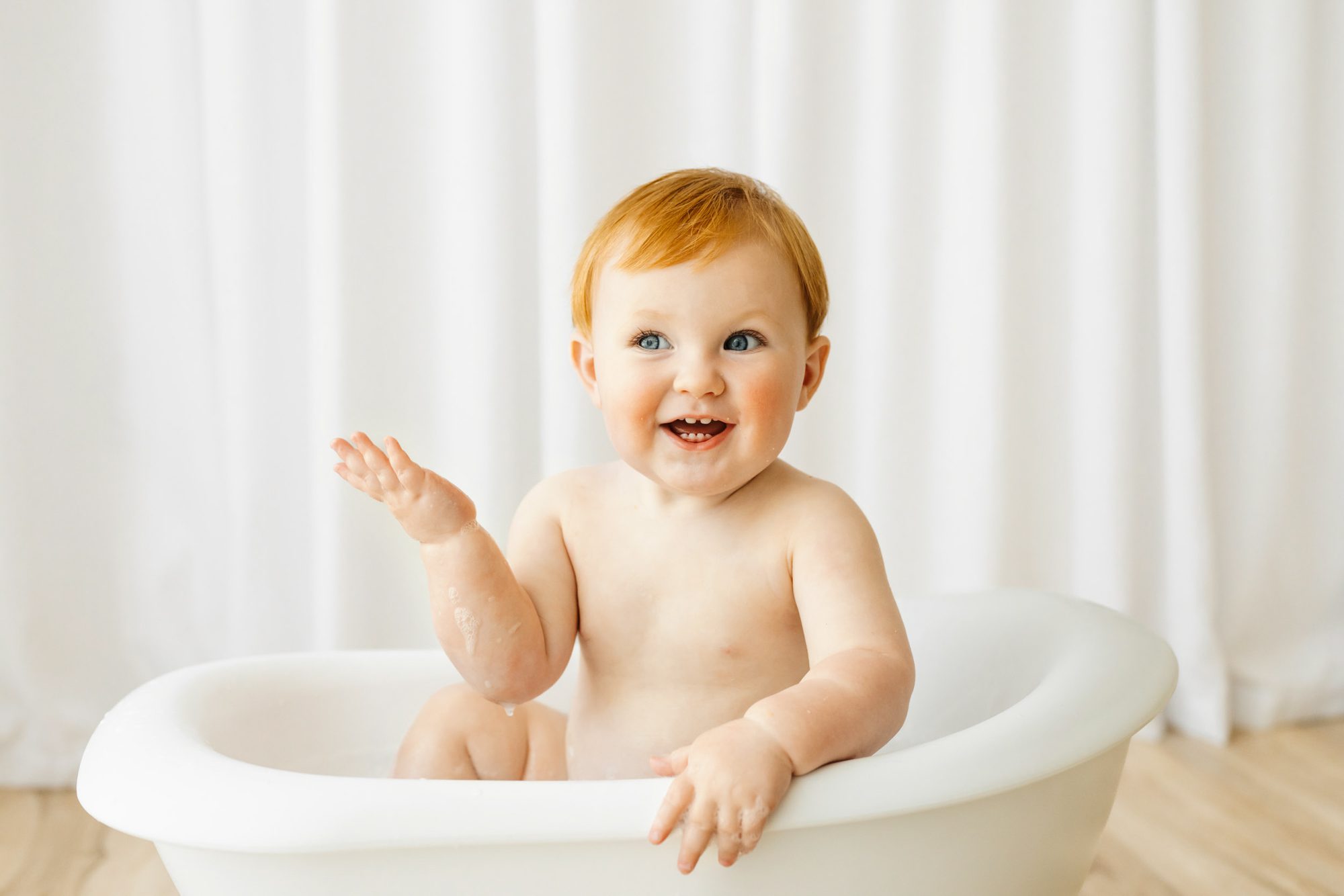 A NAPCP master baby photographer portfolio image of a little boy in a small white bathtub looking off to the side with a toothy smile and lifting his hand up in the air during a 1st birthday cake smash & splash photoshoot