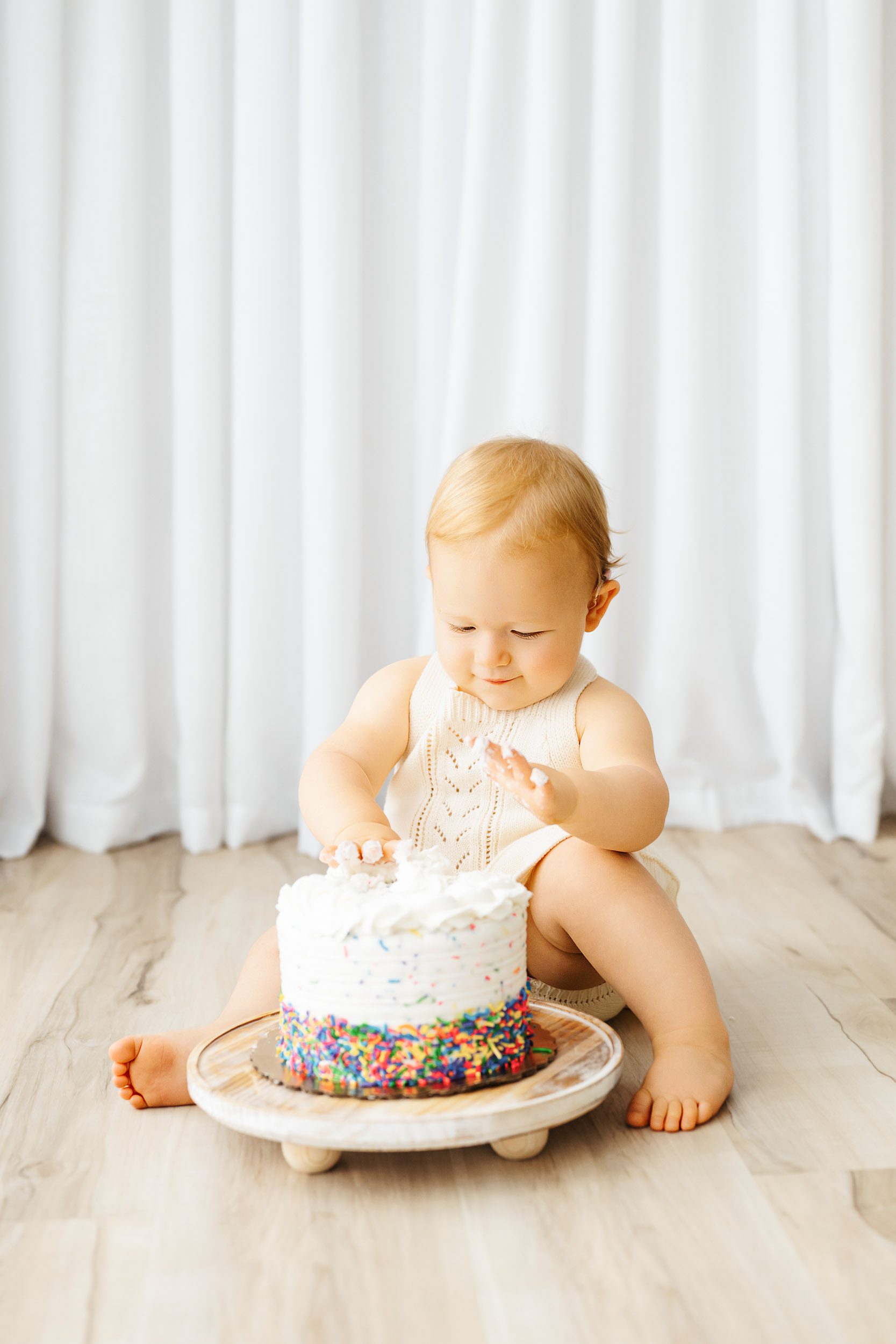 a young girl sitting on the floor and playing with the icing on her birthday cake during a cake smash photoshoot