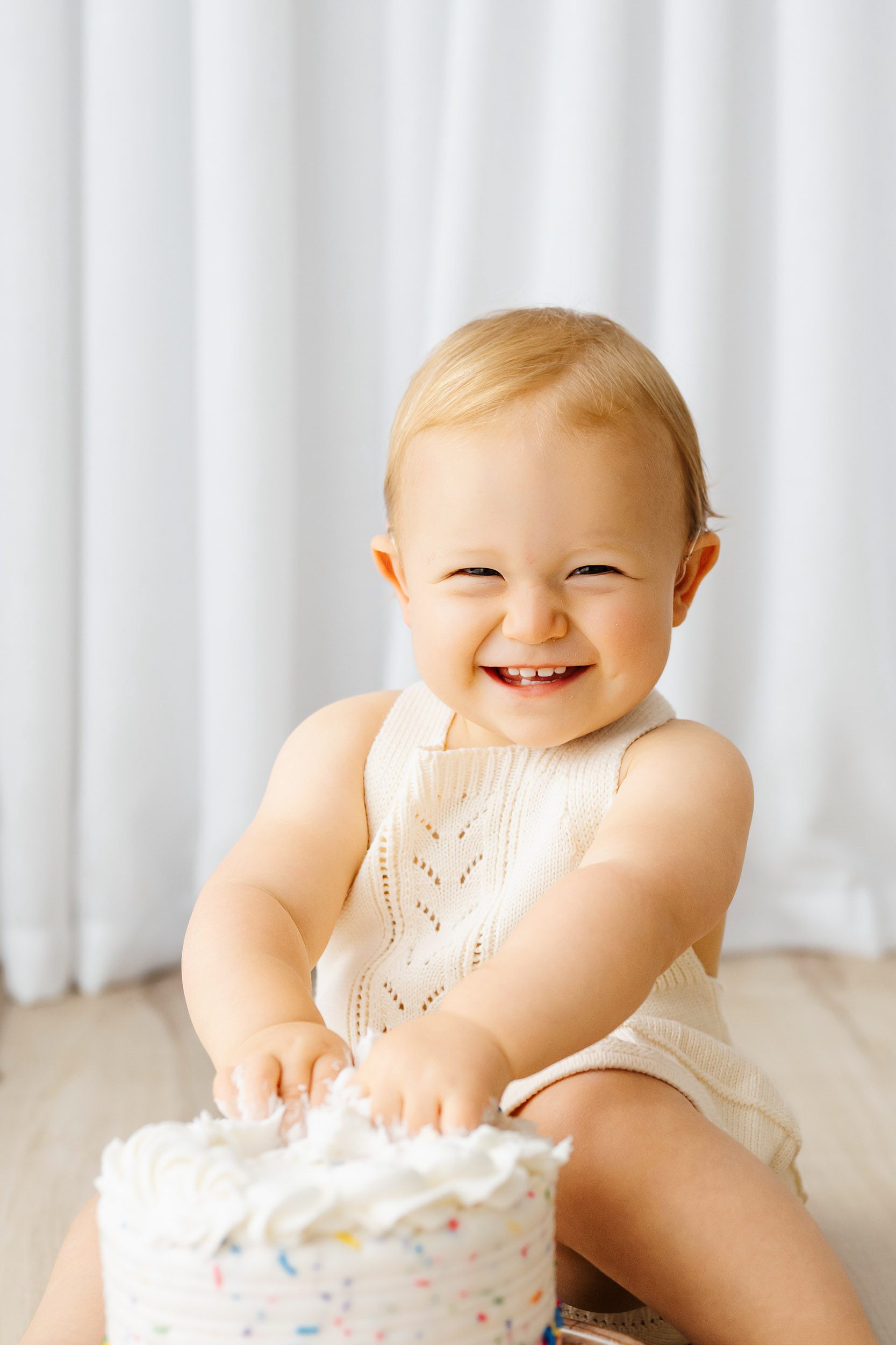 a little girl playing with the icing on her birthday cake as she looks at the camera with a huge smile on her face during a 1st birthday cake smash photoshoot