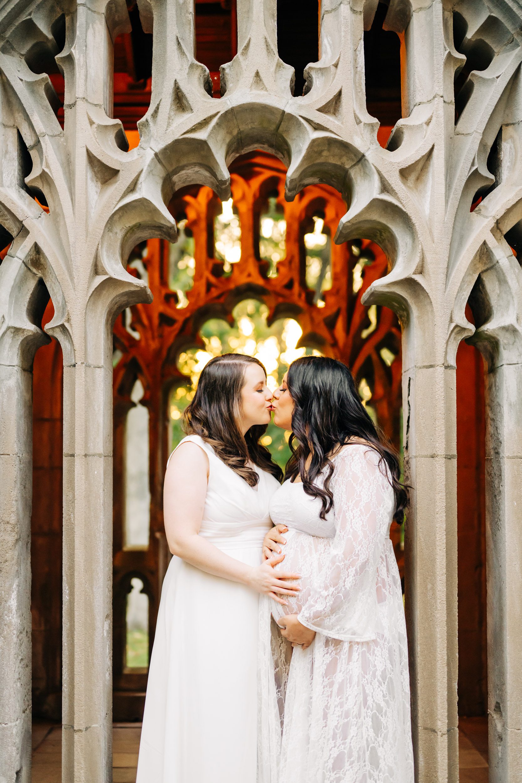 an expecting mother and her wife standing in a stone archway and kissing each other as they both rest their hands on mom's belly during an LGBT maternity photo session