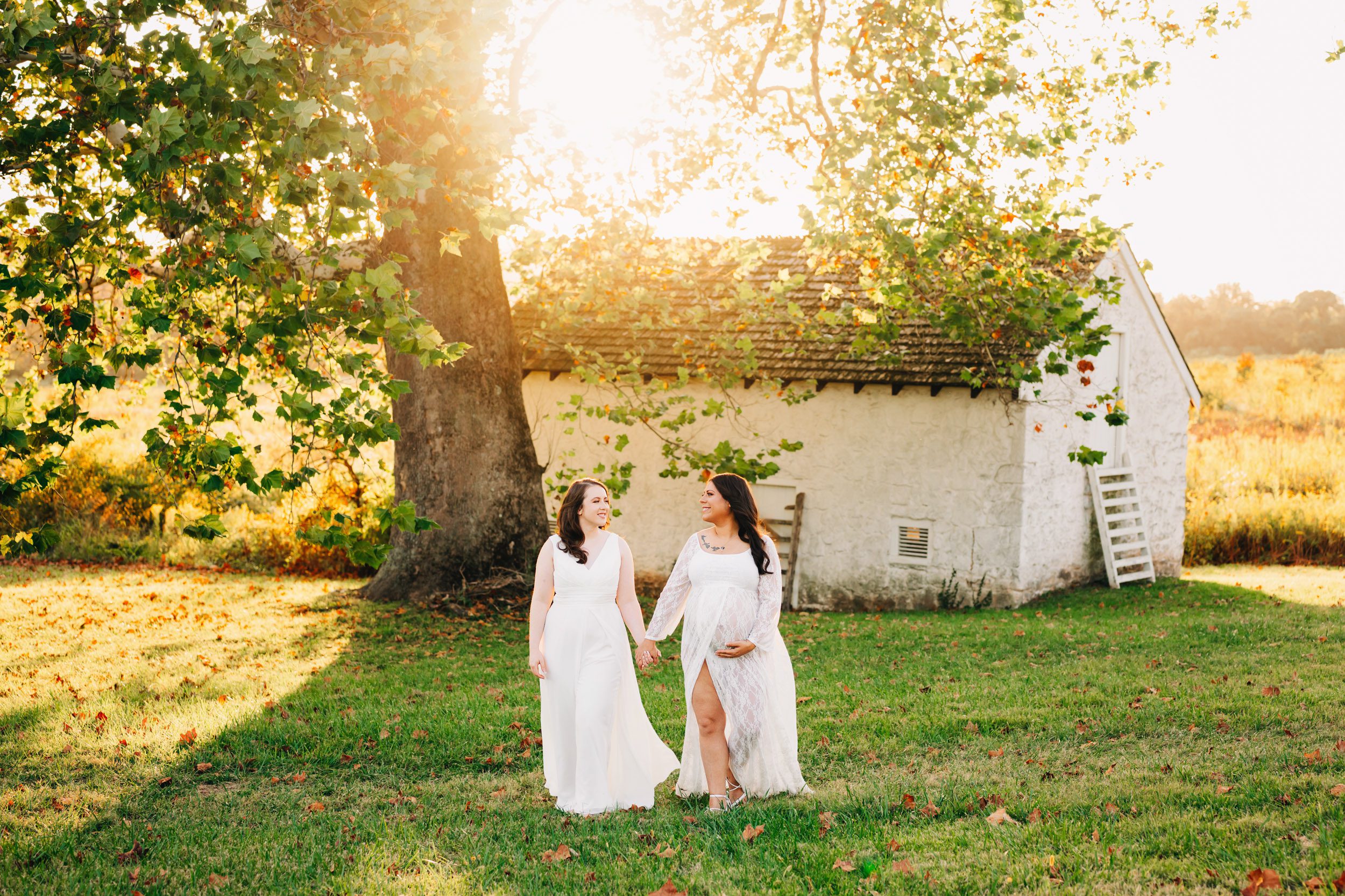 an expecting mom and her wife walking through the grass holding hands as they smile at each other with the sunset shining through the leaves in the background during an LGBT maternity photo session