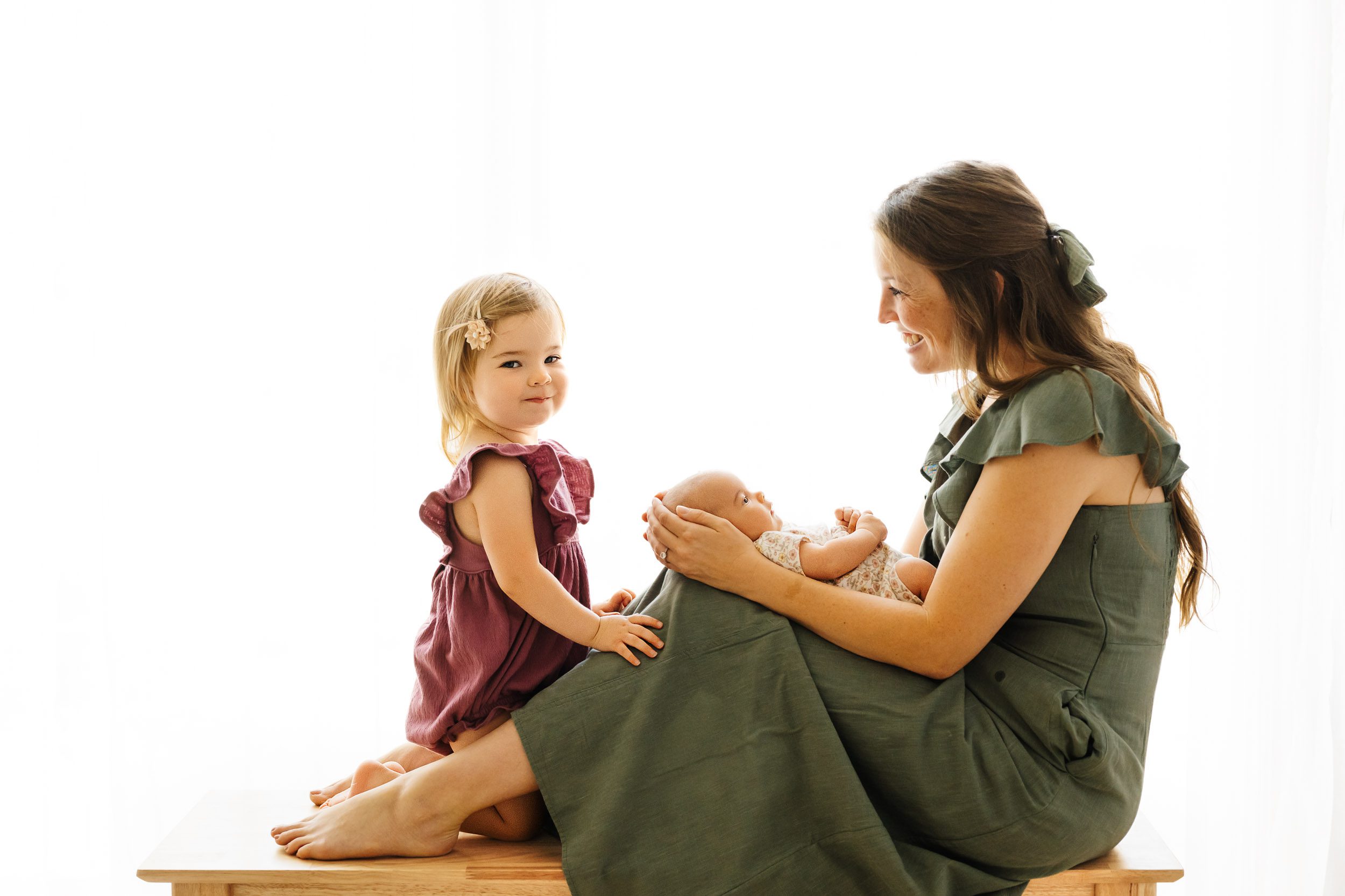 a backlit photo of a mom sitting on a bench and holding her baby girl on her lap and smiling at her older daughter as she looks at the camera during a baby photography session