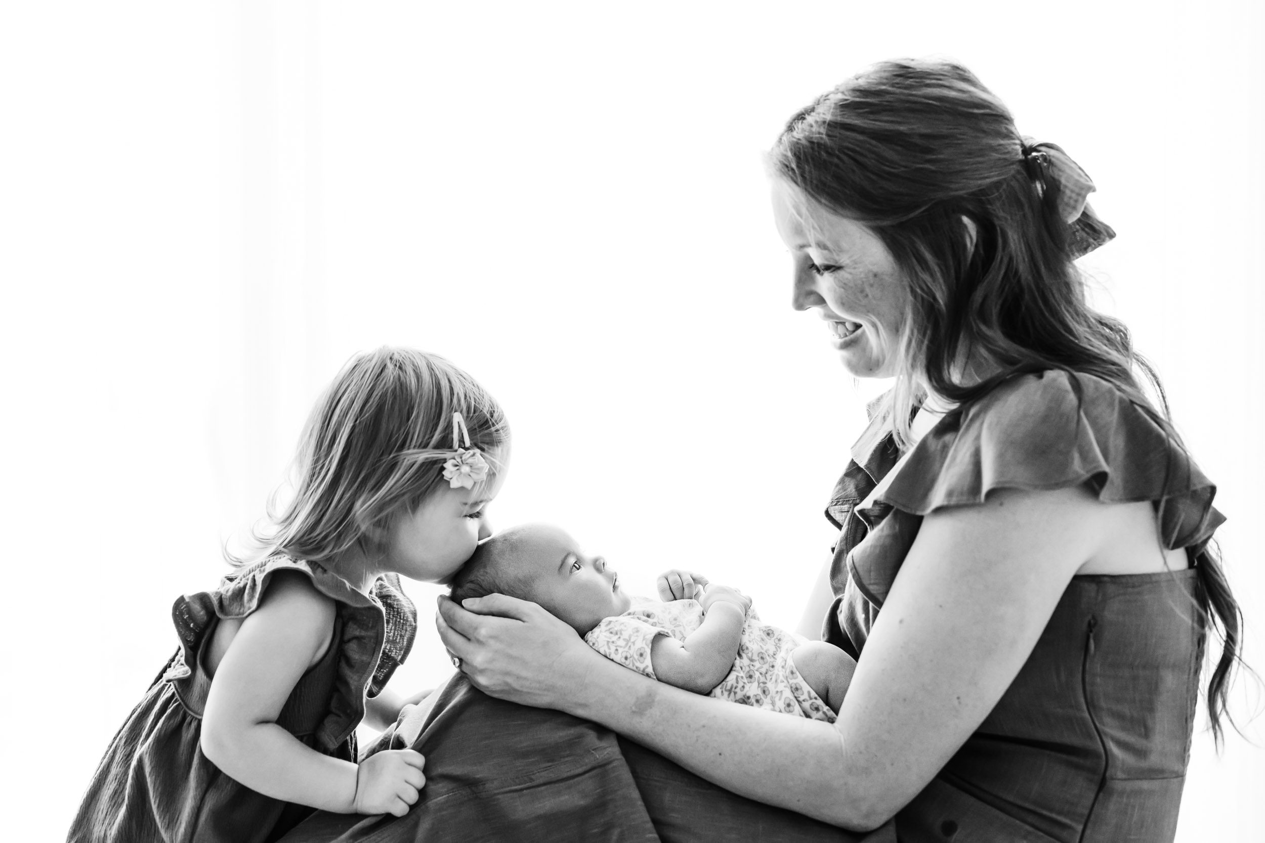 a black and white backlit photo of a mom holding her baby girl on her lap as big sister leans in and gently kisses her baby sister on the head during a baby photography session