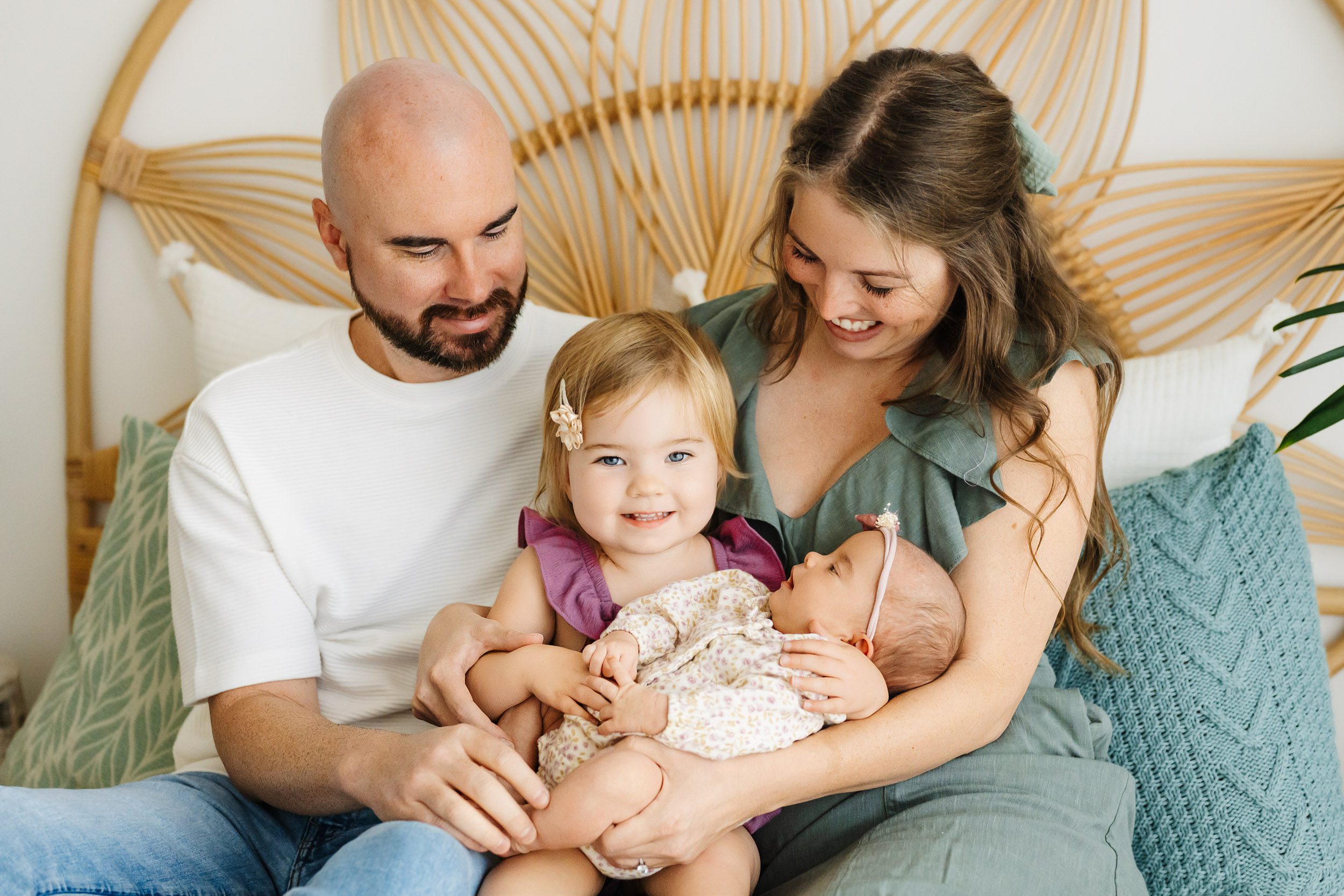 a family of four sitting on a bed with big sister holding her baby sister on her lap and looking at the camera with a big smile on her face as mom and dad smile down at the girls during a baby photography session