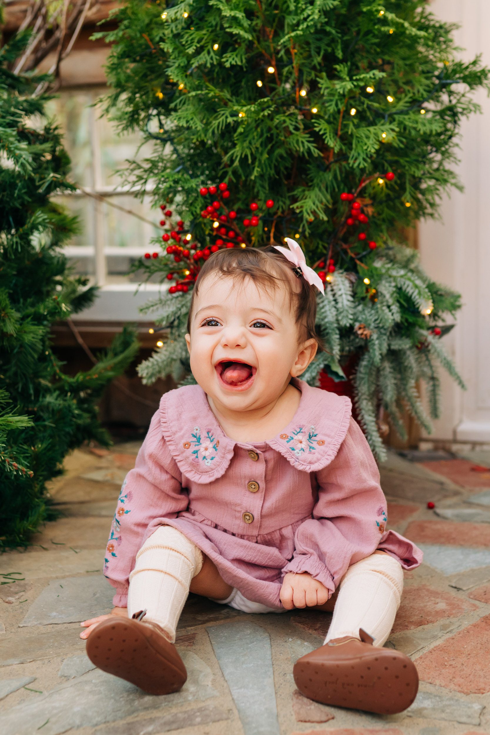 a baby girl sitting on the floor in front of a tree decorated with Christmas lights and red berries as she smiles and sticks her tongue out during a holiday mini session