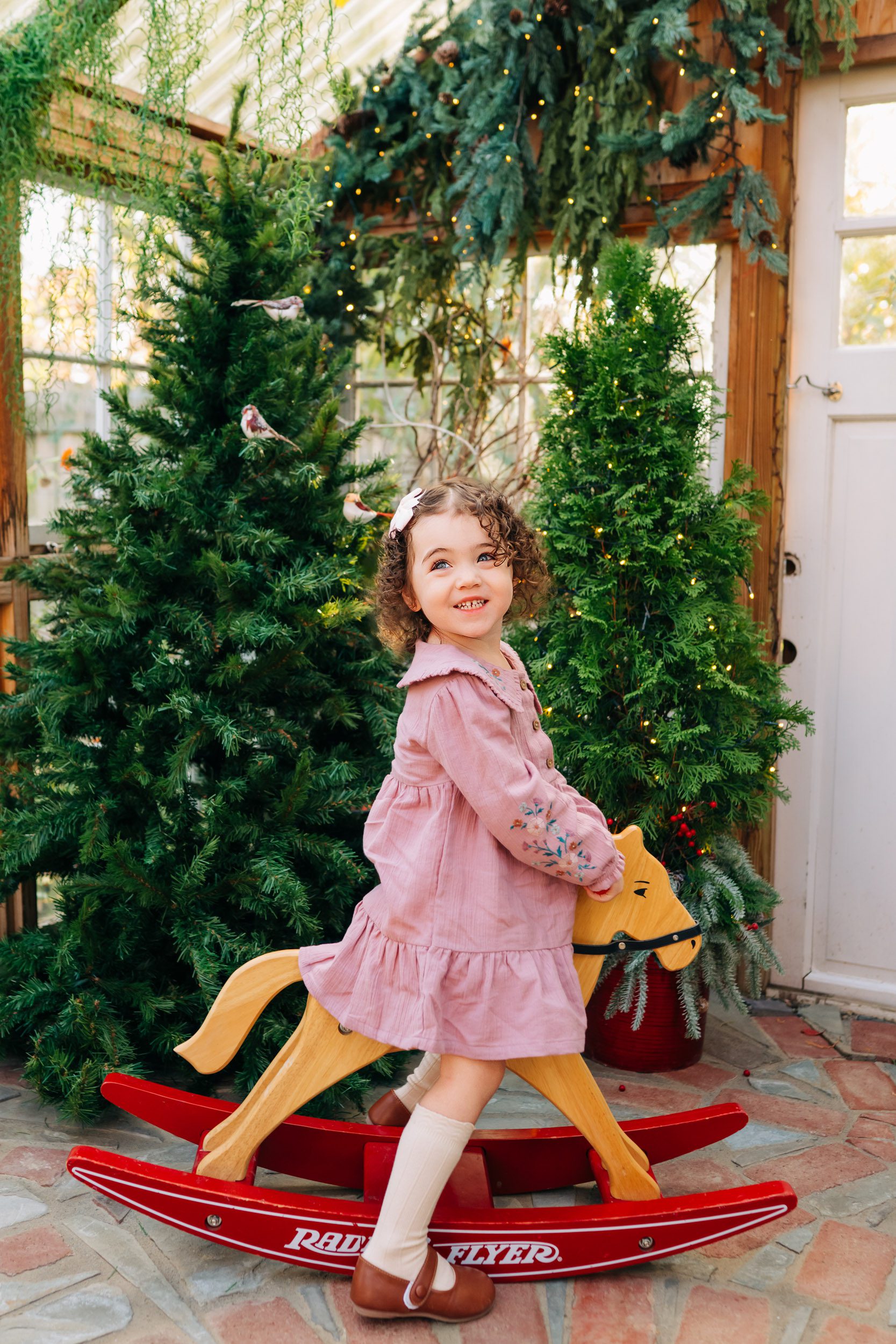 a young girl riding a rocking horse with Christmas trees in the background during a holiday mini session