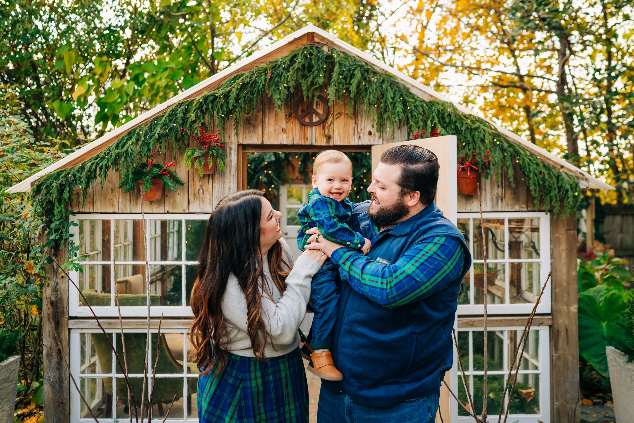 parents standing in front of a small greenhouse decorated with holiday decor and holding their young son in their arms and tickling him as he smiles at the camera during a holiday mini session