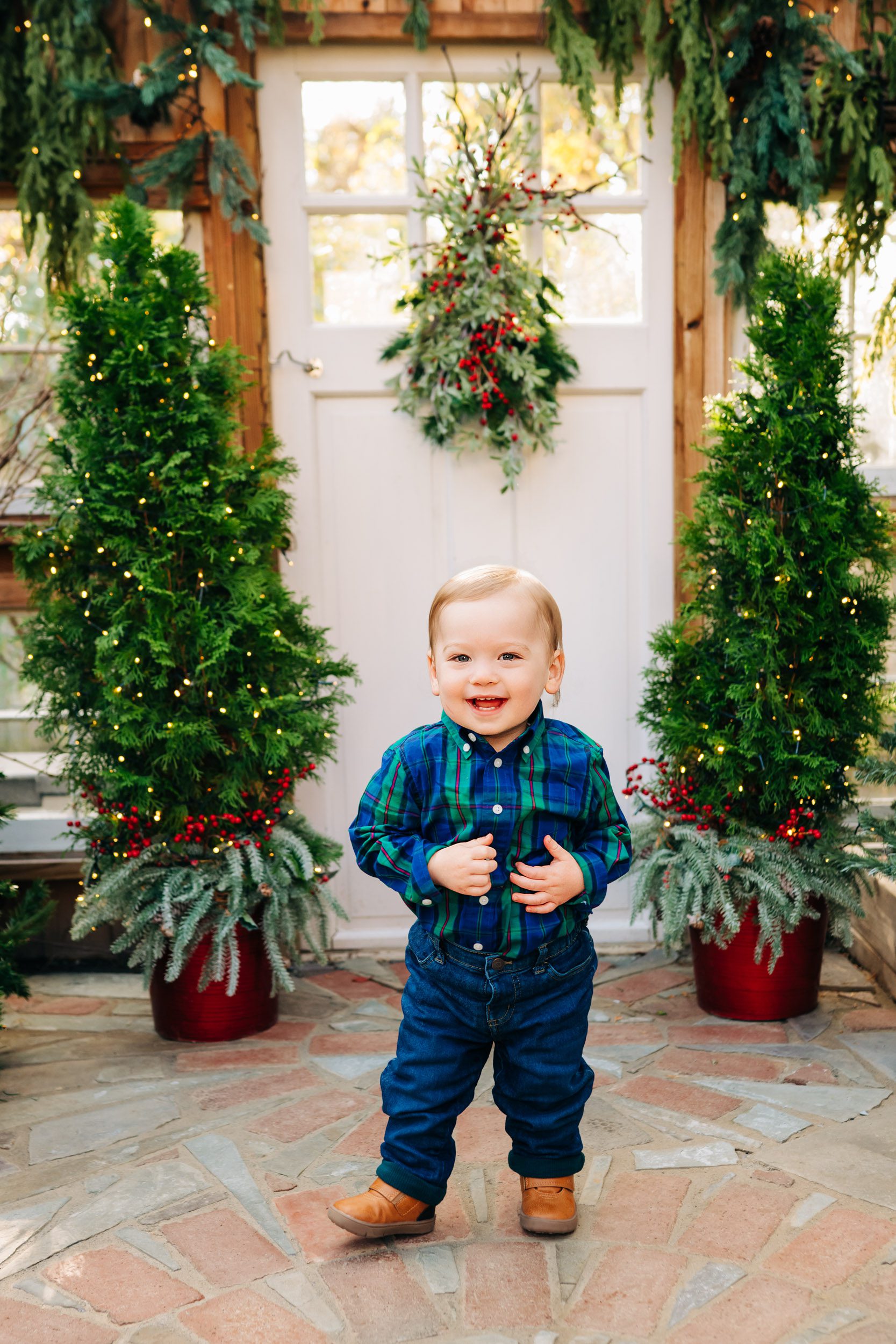 a little boy wearing a blue and green plaid shirt standing in a greenhouse surrounded by Christmas trees and holiday decor as he smiles at the camera during a holiday mini session