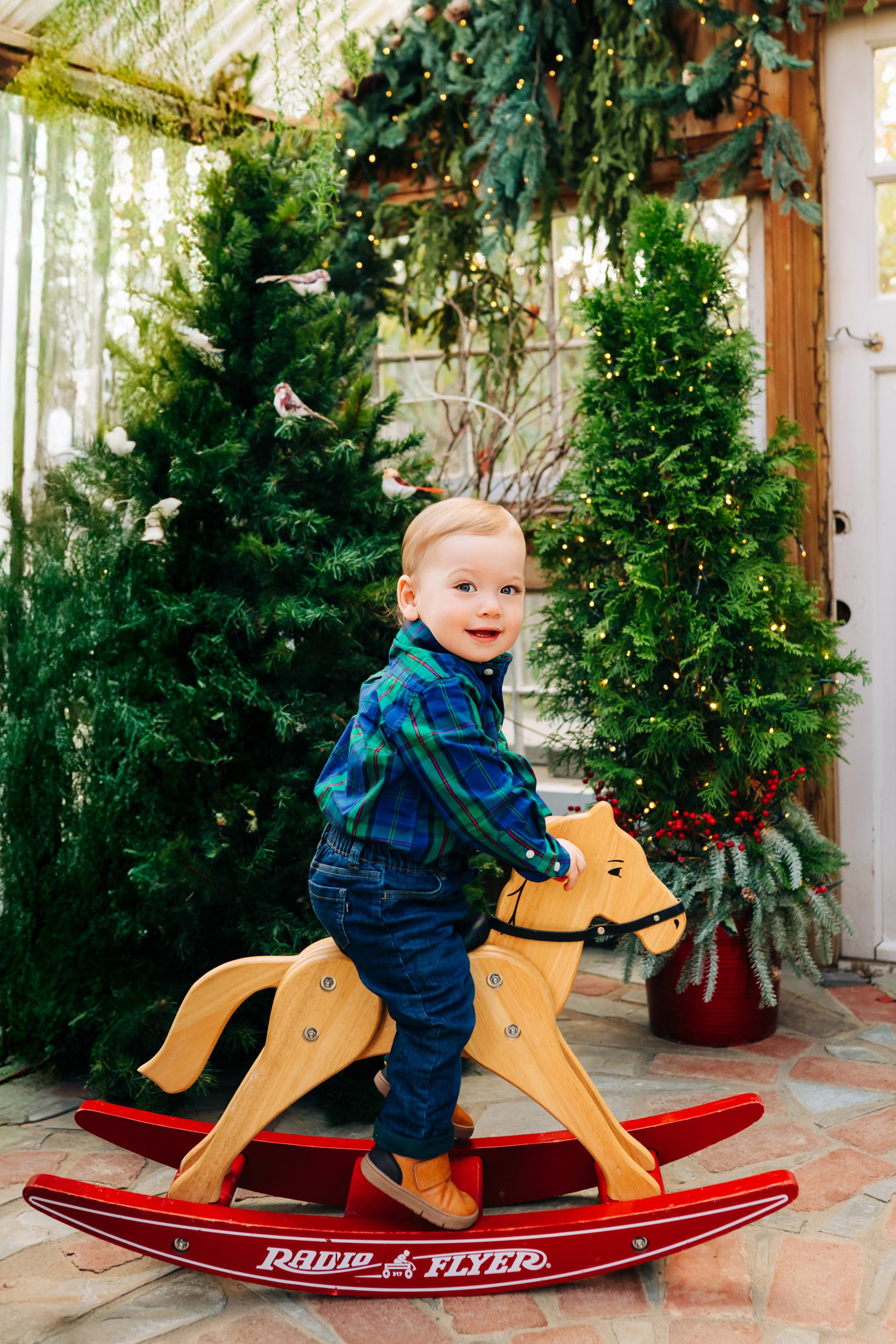 a young boy riding a rocking horse with Christmas trees in the background during a holiday mini session