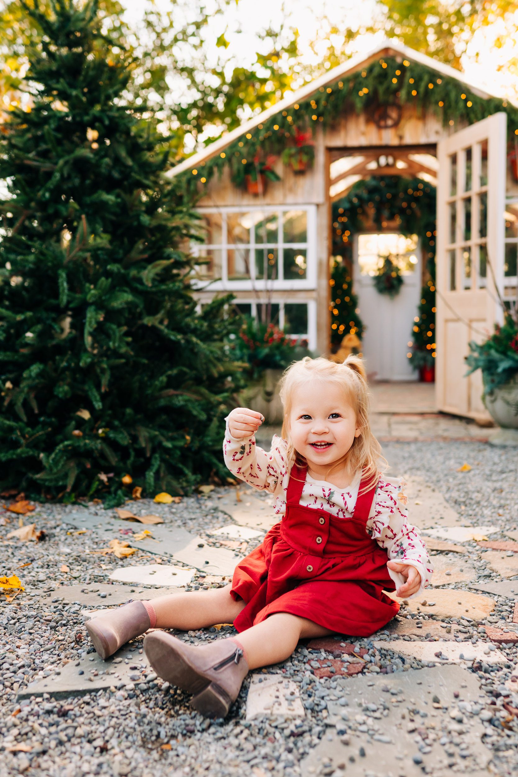 a young girl wearing a red jumper sitting on the ground in front of a small greenhouse and playing with the stones with a huge smile on her face during a holiday mini session