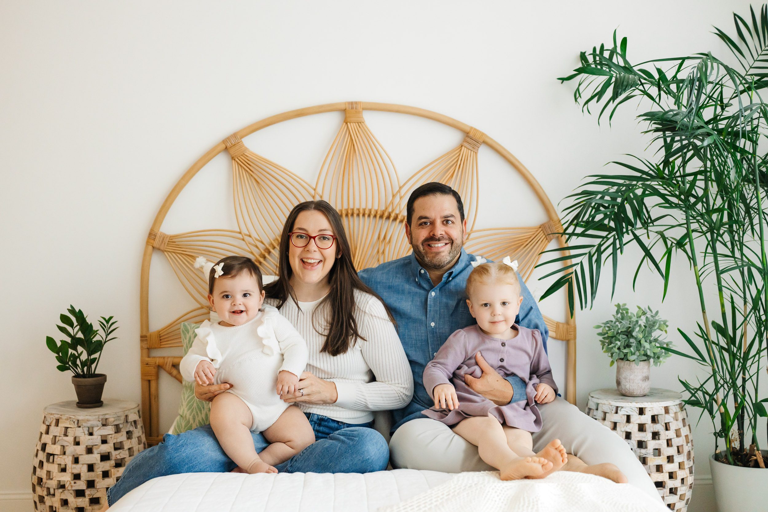 a family of four sitting on a bed together with each parent holding one of their young daughters on their lap as they all look at the camera and smile during a one year old photoshoot