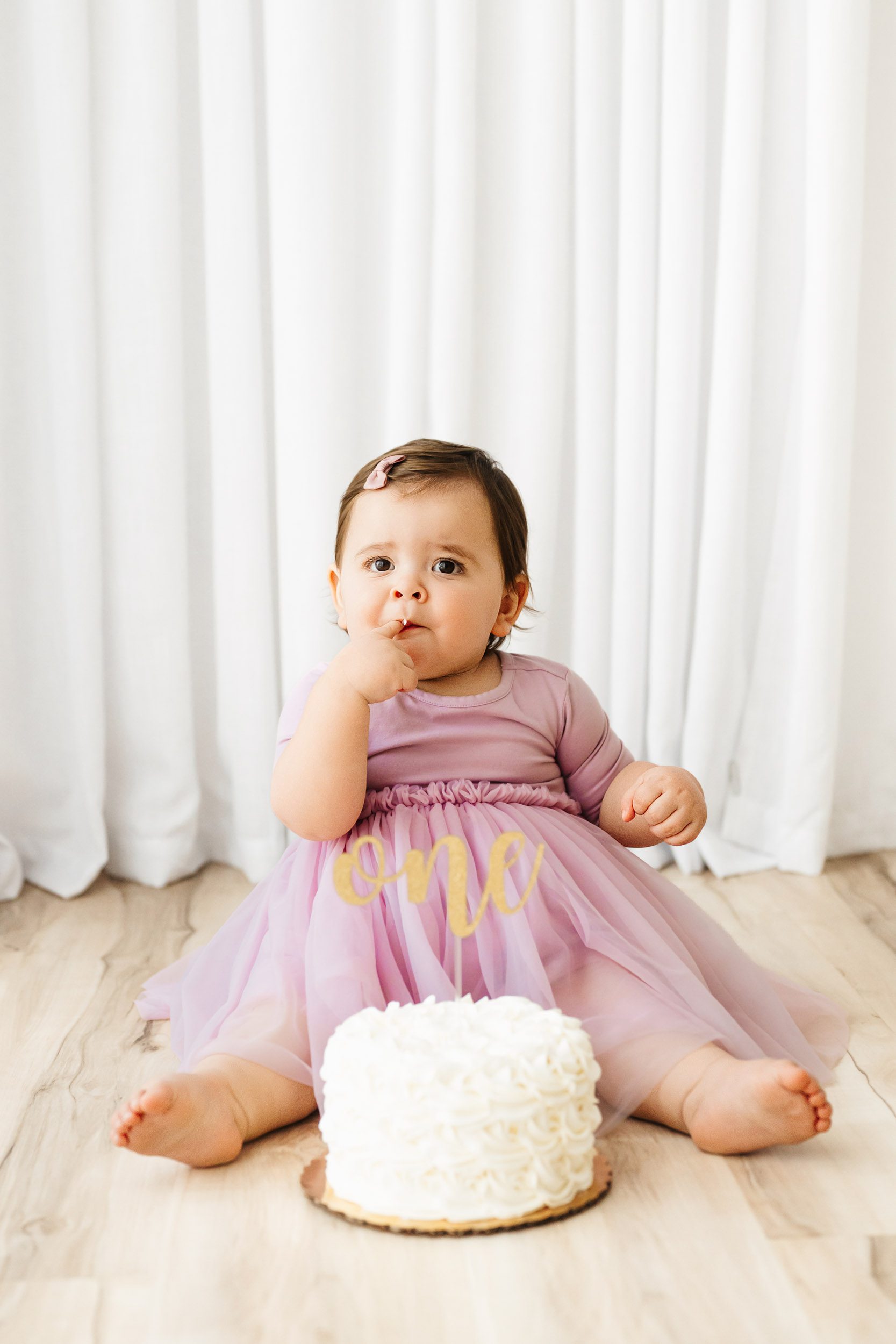 a little girl sitting on the floor with her first birthday cake in front of her and licking icing off one of her fingers during a one year old photoshoot