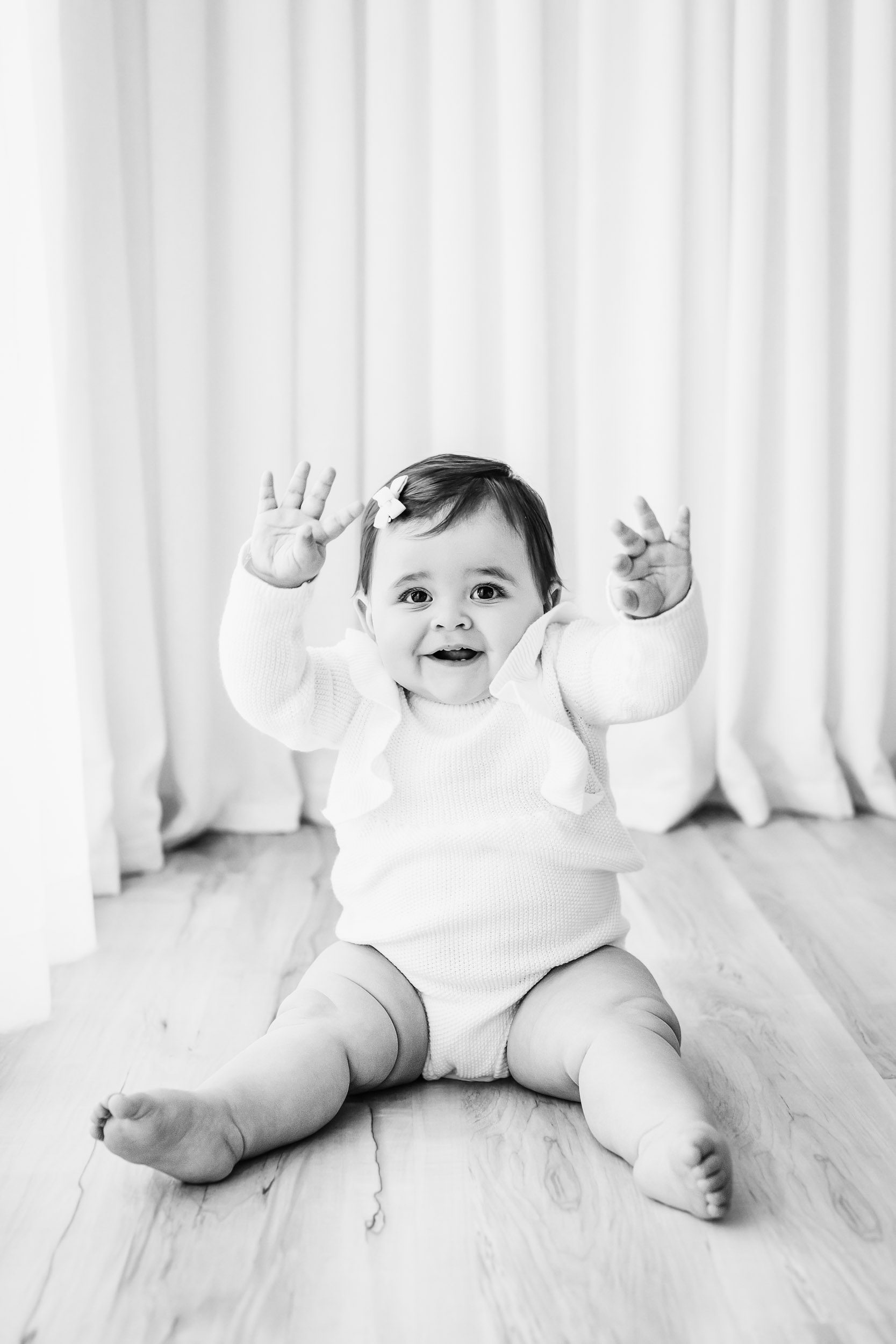 a black and white photo of a young girl wearing a white romper lifting her hands in the air and smiling at the camera during a one year old photoshoot