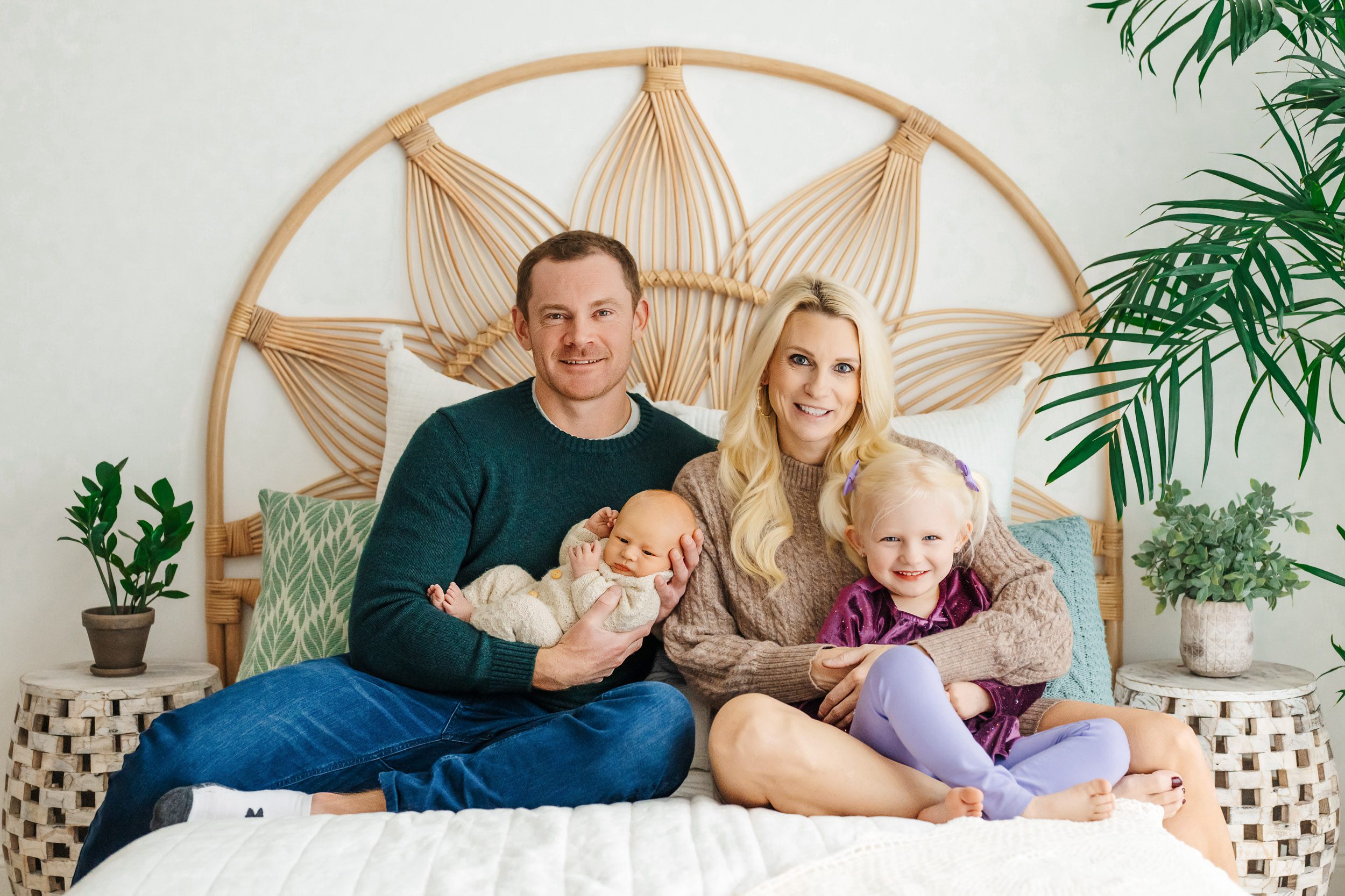 a family of four sitting on a bed together with dad cradling his baby boy in his arms and mom holding her older daughter on her lap as they all smile at the camera during a newborn photography session