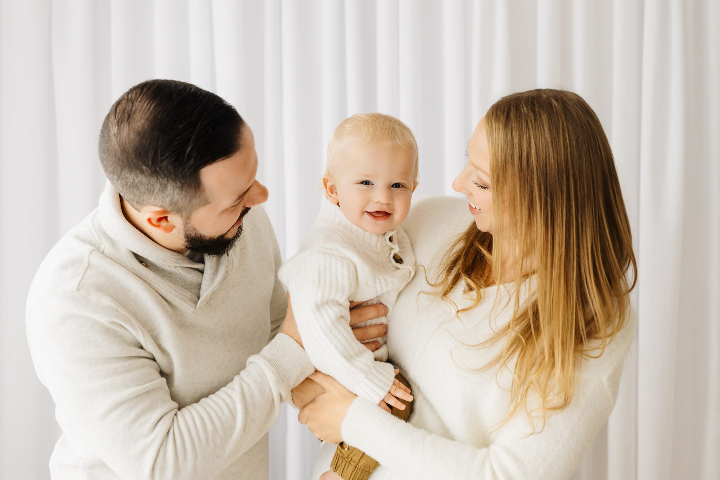 parents standing in front of a wall of white curtains holding their young son in their arms and smiling at him as he smiles directly at the camera during his 1st birthday pictures
