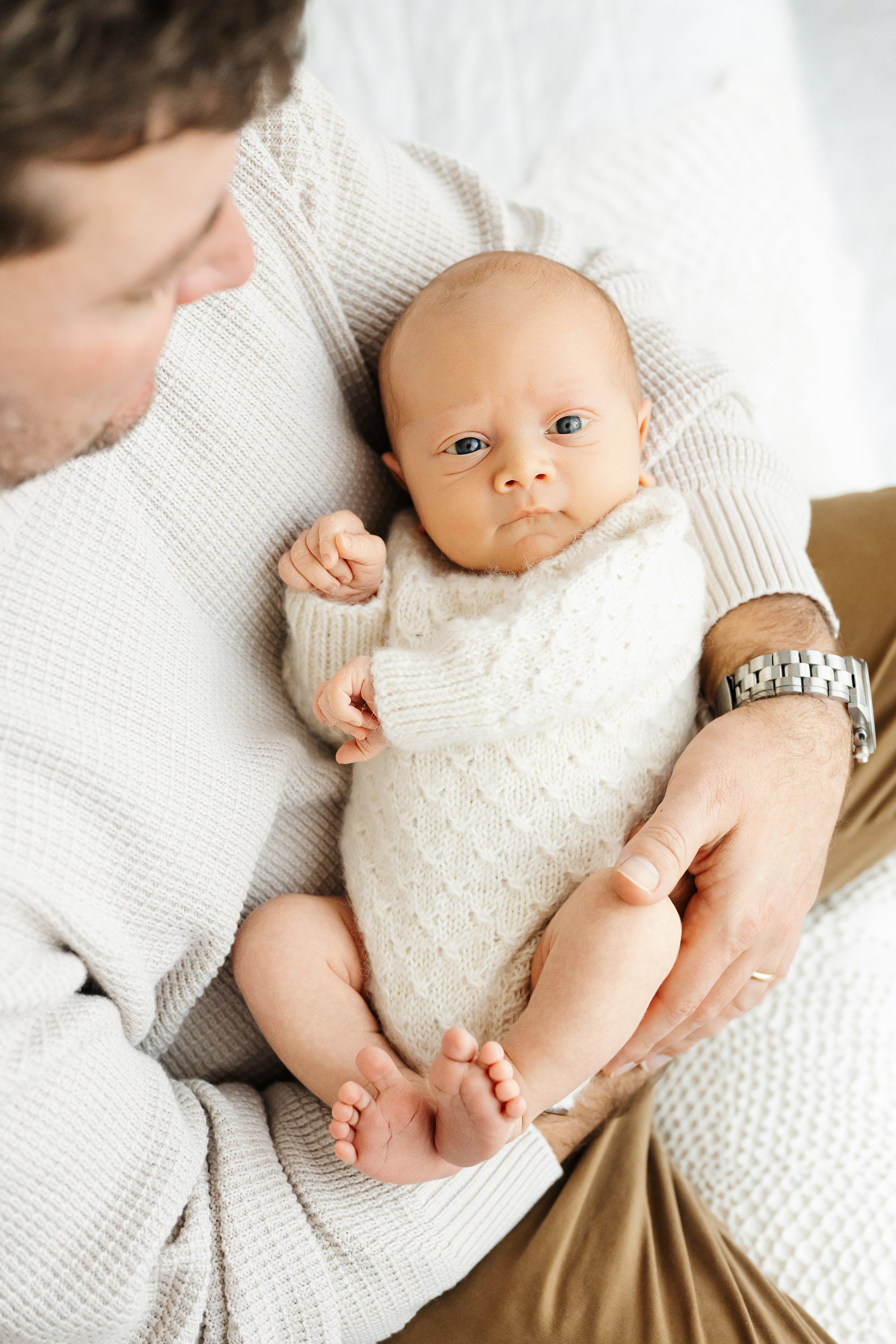 a picture taken from above of a baby boy wearing a long sleeve textured knit romper snuggled in his dad's arms and gazing up at the camera during a natural newborn photoshoot