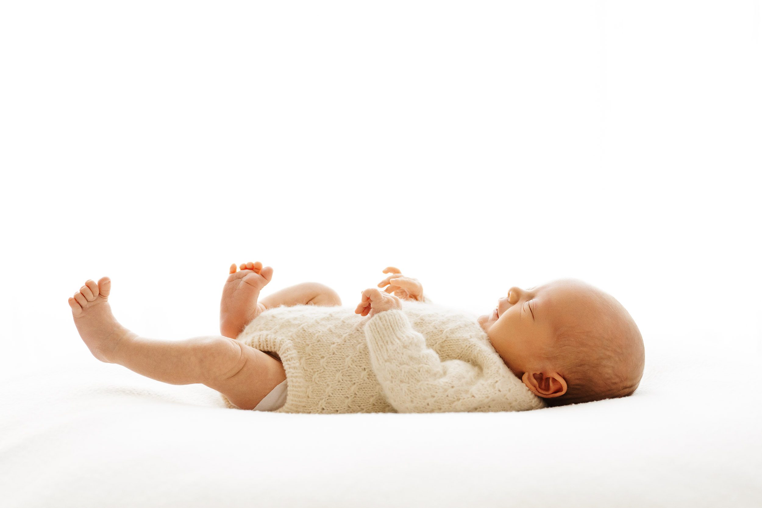 a backlit photo of a baby laying on his back and kicking his legs with a hint of a smile on his face during a natural newborn photoshoot