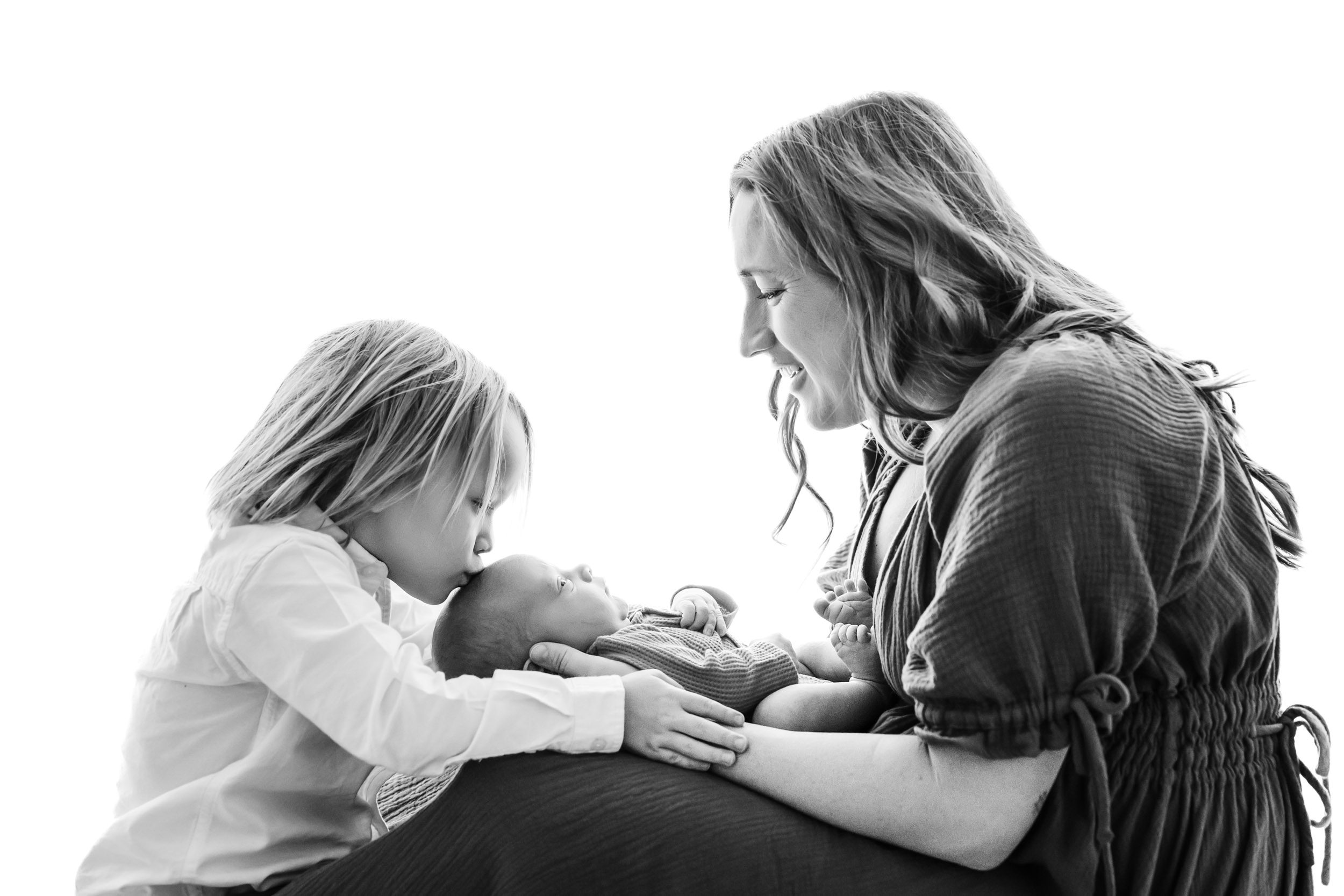 a black and white backlit photo of a mom holding her baby boy in her arms as her older son gives his baby brother a kiss on the head during a lifestyle newborn photoshoot
