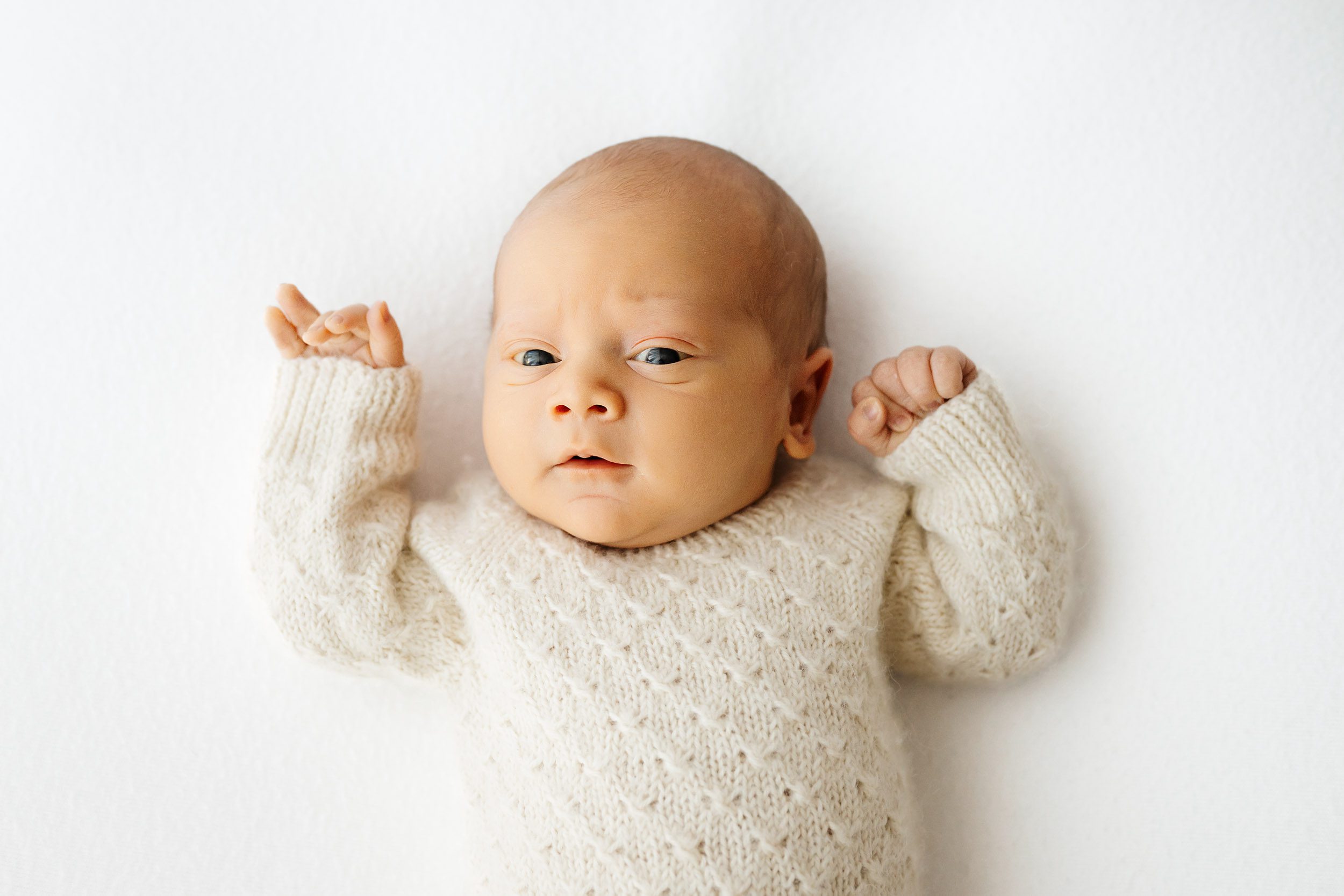 a baby boy wearing a long sleeve textured knit romper holds his hands up next to his face and gazes directly at the camera during a lifestyle newborn photoshoot