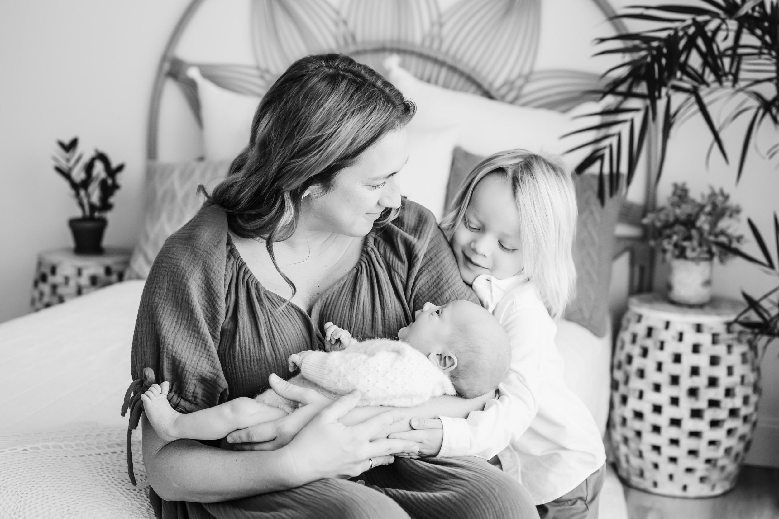 a black and white photo of a mom sitting on a bed holding her newborn son in her arms as her older son hugs her and smiles down at his baby brother during a lifestyle newborn photoshoot