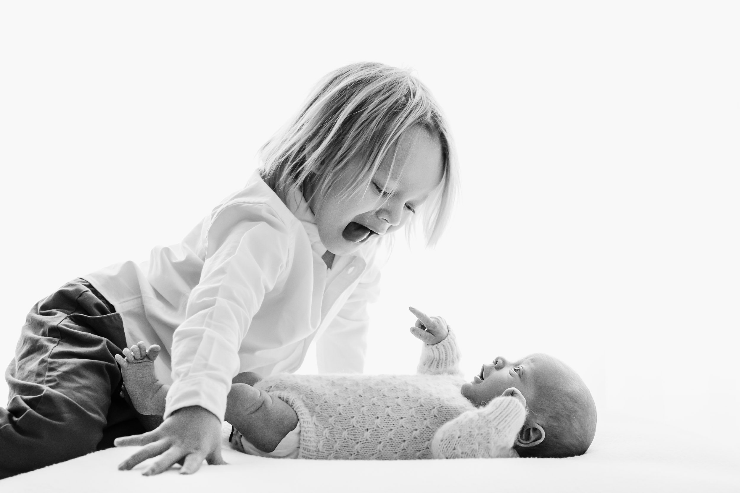 a black and white backlit photo of a baby laying on a bean bag and pointing up at his big brother, who is looking down at his baby brother and sticking his tongue out during a natural newborn photoshoot
