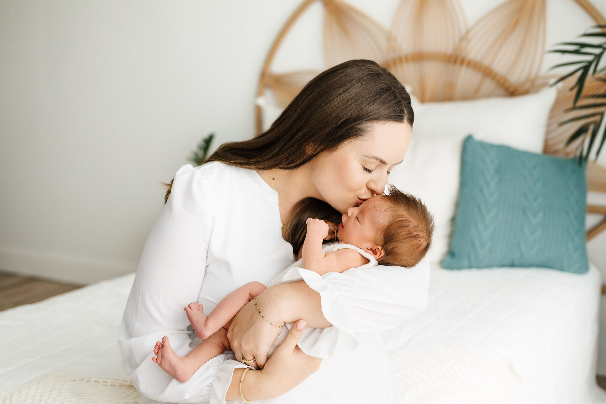 a new mom sitting on a bed and cradling her newborn baby girl in her arms as she gently kisses the baby on the forehead during a lifestyle newborn photoshoot