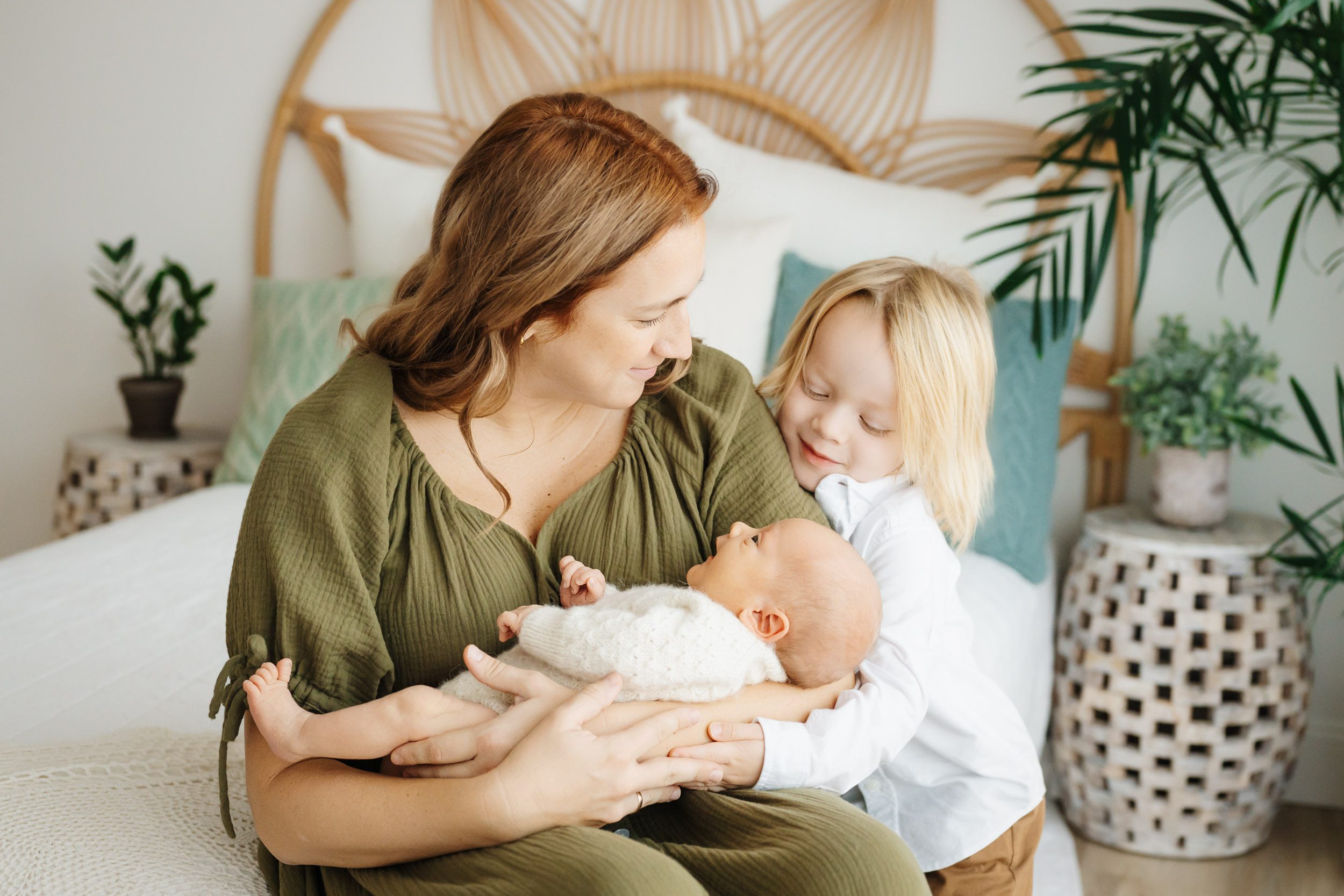 a new mom sitting on a bed cradling her baby boy in her arms as her older son hugs his mom and smiles down at the baby during a lifestyle newborn photoshoot
