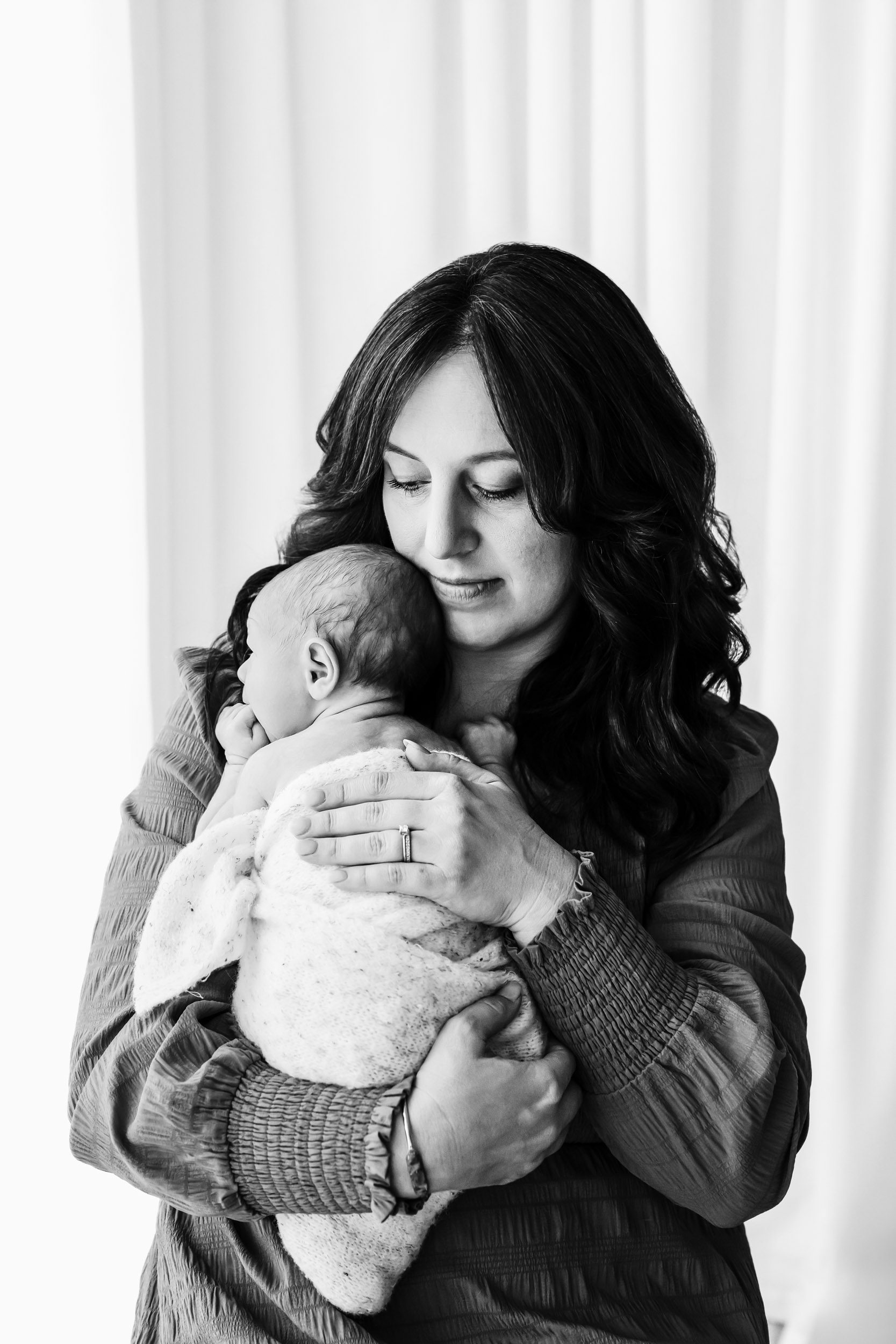 a black and white picture of a new mom holding her baby closer to her chest and resting her chin on his head during a lifestyle newborn photoshoot