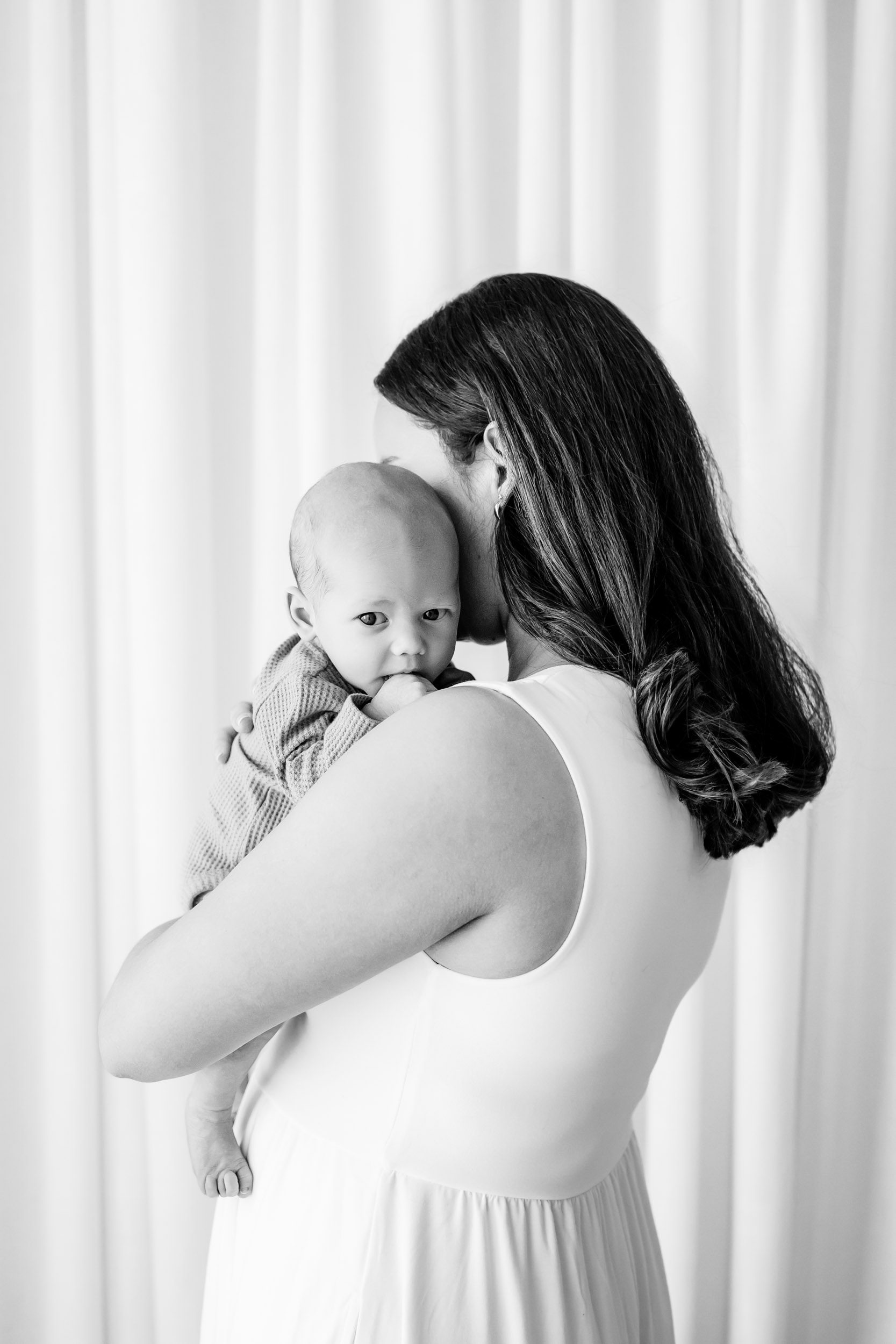 a black and white picture of a new mom holding her baby boy cuddled up against her chest as he looks over her shoulder at the camera during a lifestyle newborn photoshoot