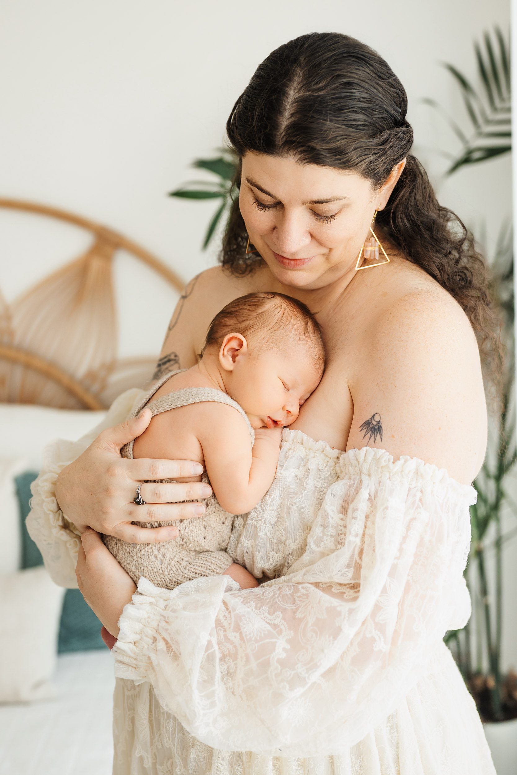 a new mom wearing a lacy white off the shoulder dress holding her baby boy snuggled up against her chest during a newborn photoshoot