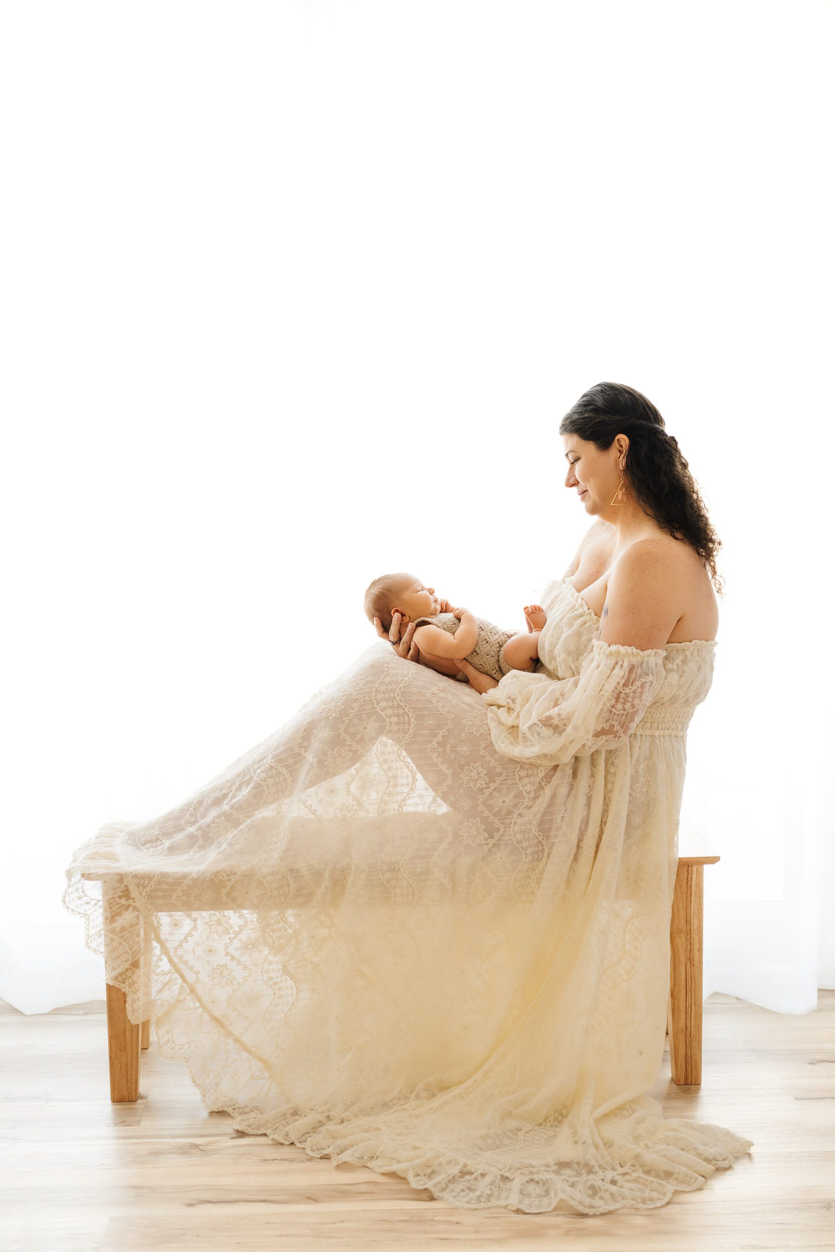 a backlit photo of a new mom wearing a lacy white dress sitting on a bench holding her baby boy and smiling down at him during a newborn photoshoot