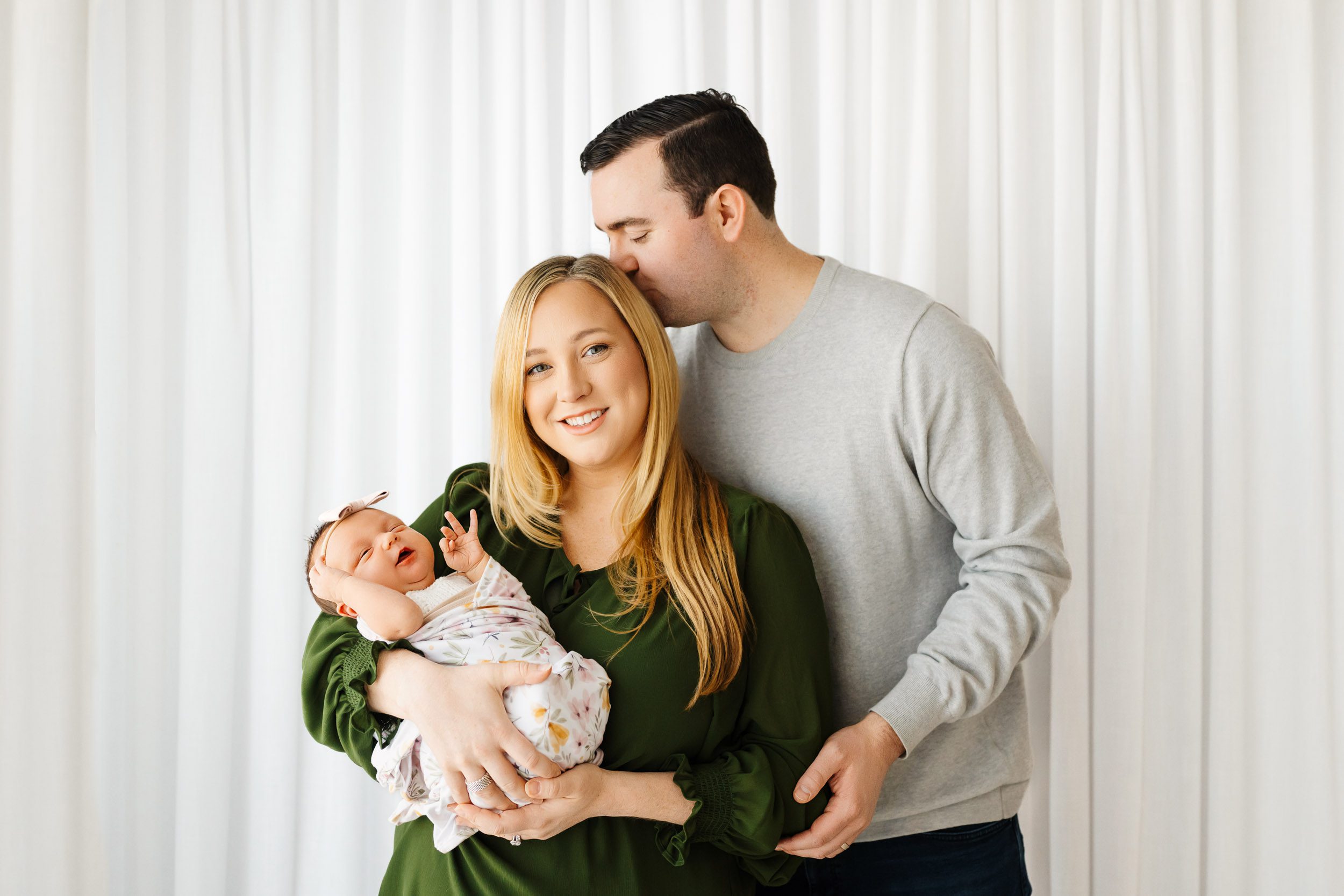 new parents standing in front of a wall of white curtains as mom cradles her baby girl in her arms and dad kisses mom on the temple during a newborn baby photography session