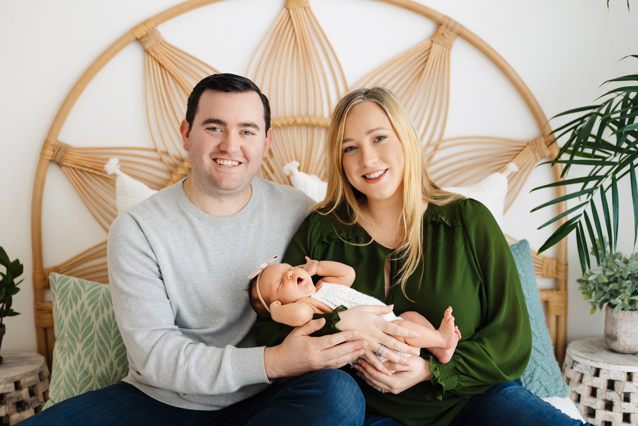 new parents sitting on a bed and smiling at the camera as they cradle their baby girl in their arms during a newborn baby photography session