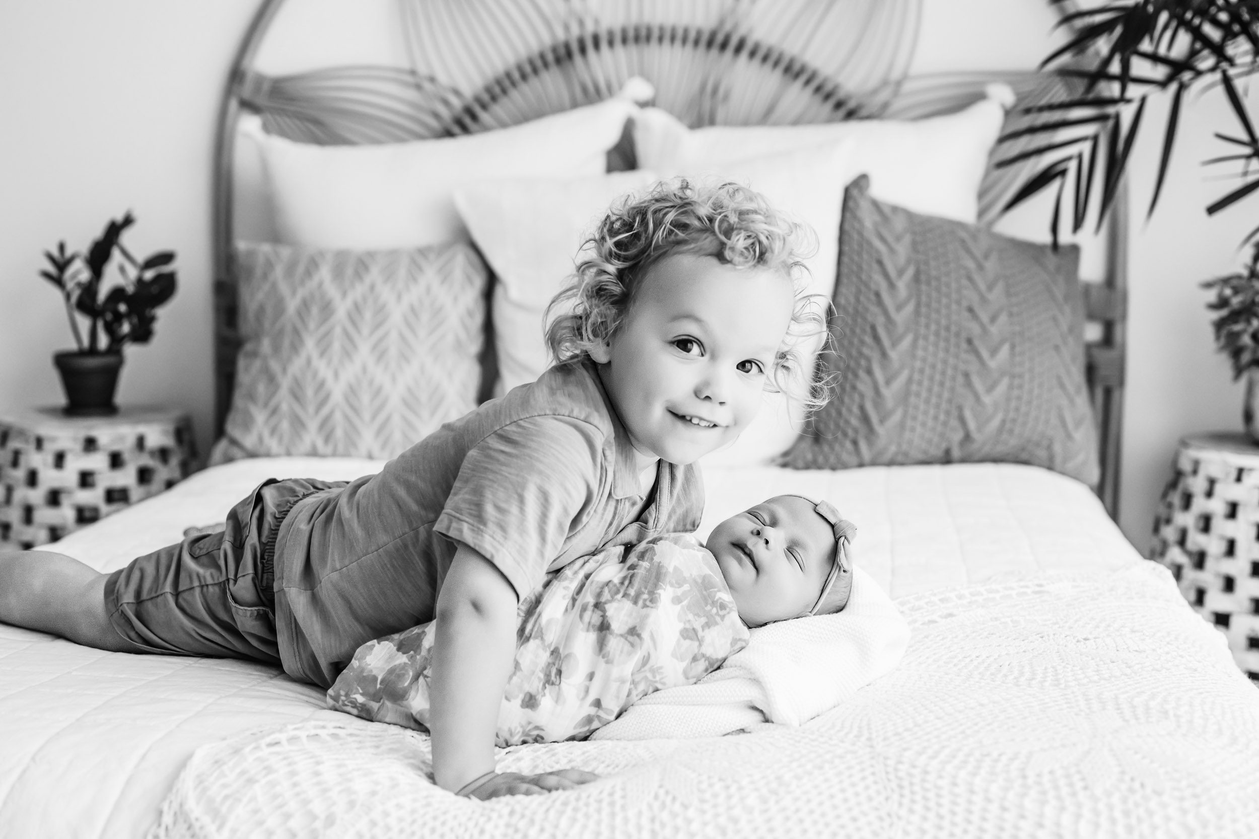 a black and white picture of a big brother laying on a bed with his baby sister as he smiles directly at the camera during a newborn photoshoot with siblings