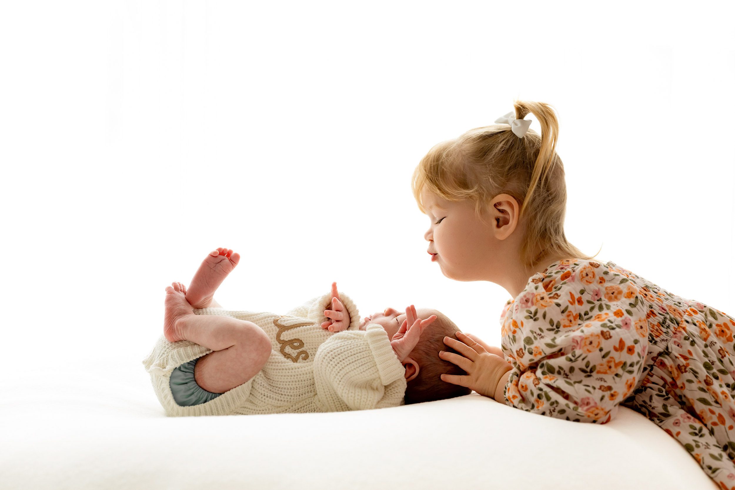 a backlit photo of a baby boy laying on a bean bag as his big sister smiles down at him during a newborn photoshoot with siblings