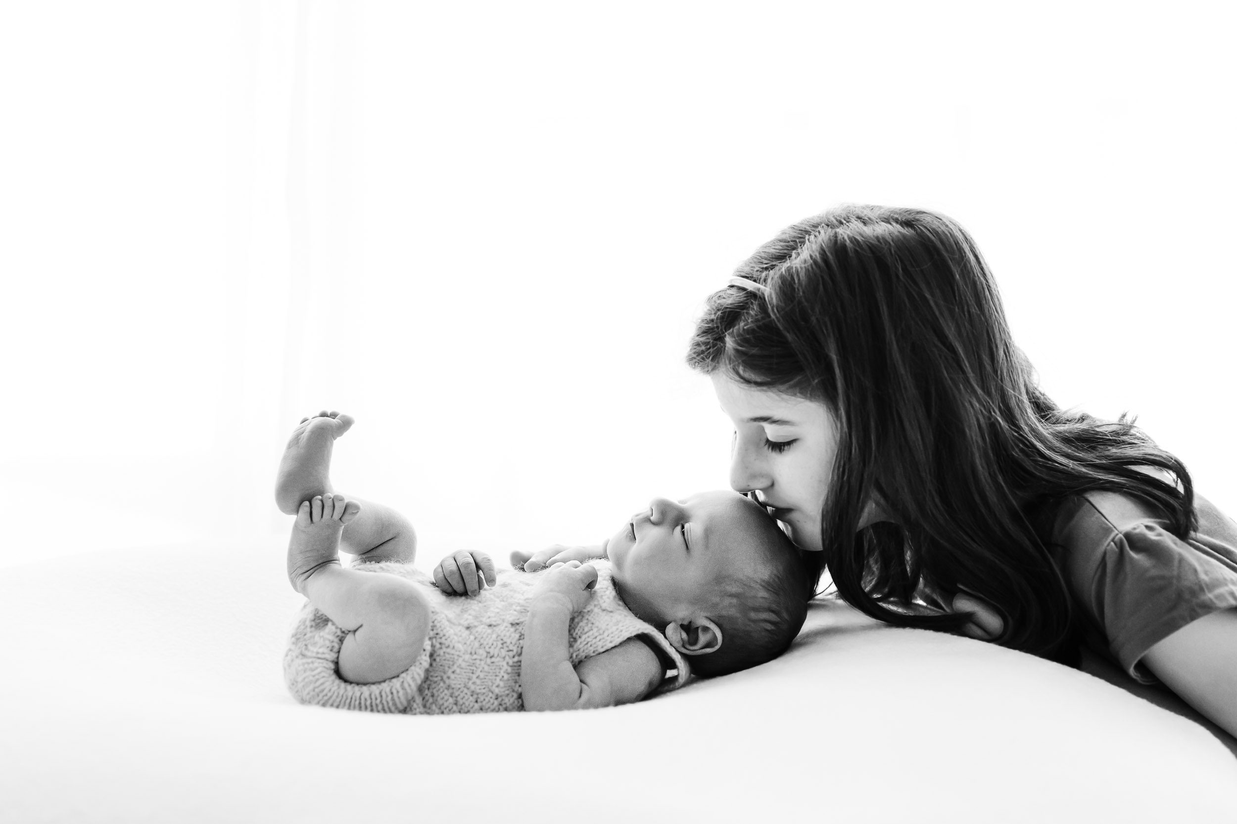 a black and white backlit photo of a baby boy laying on a bean bag as his big sister gently gives him a kiss on the head during a newborn photoshoot with siblings