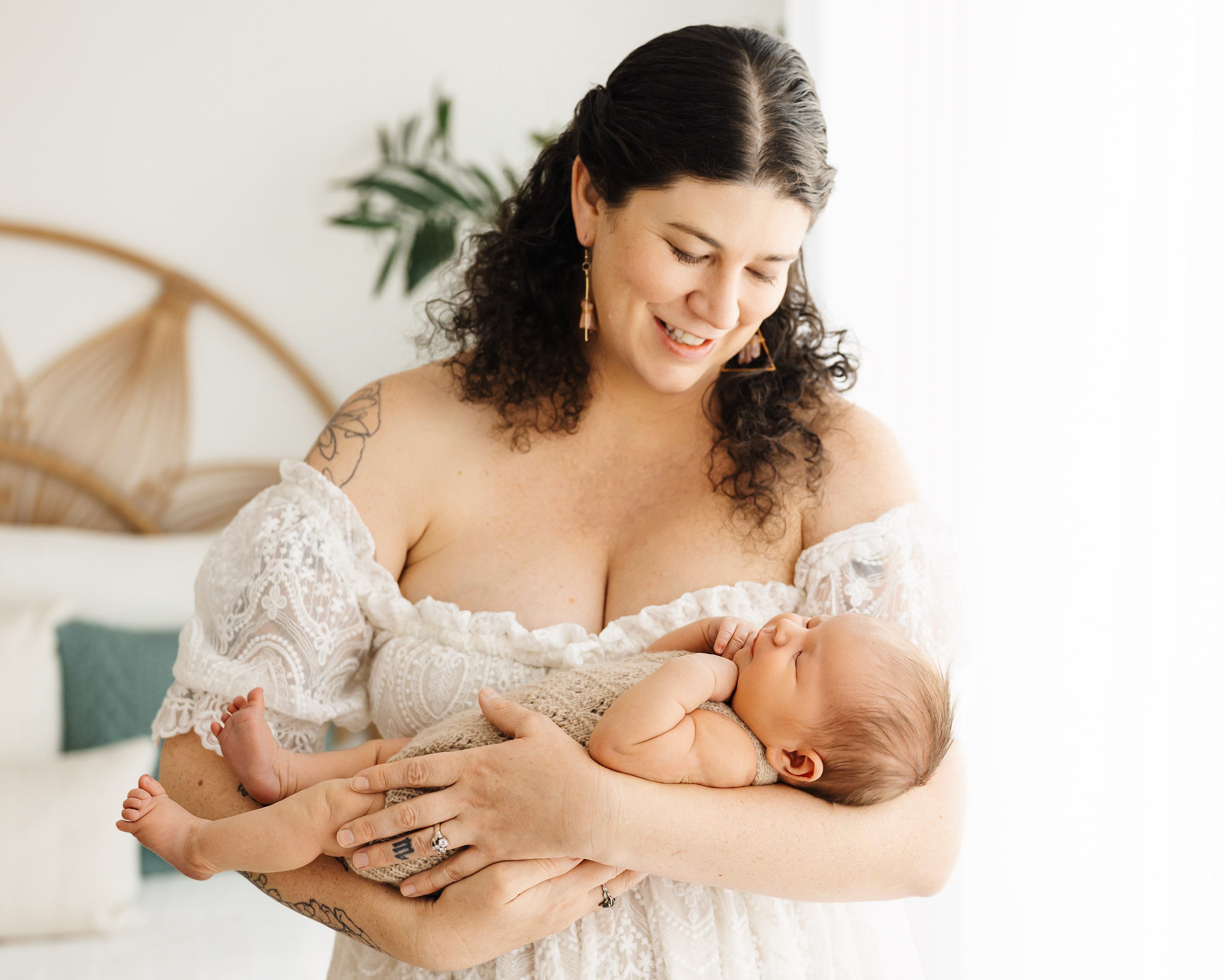 a new mom wearing a lacy white off the shoulder dress cradling her baby boy in her arms and smiling down at him during a newborn photoshoot