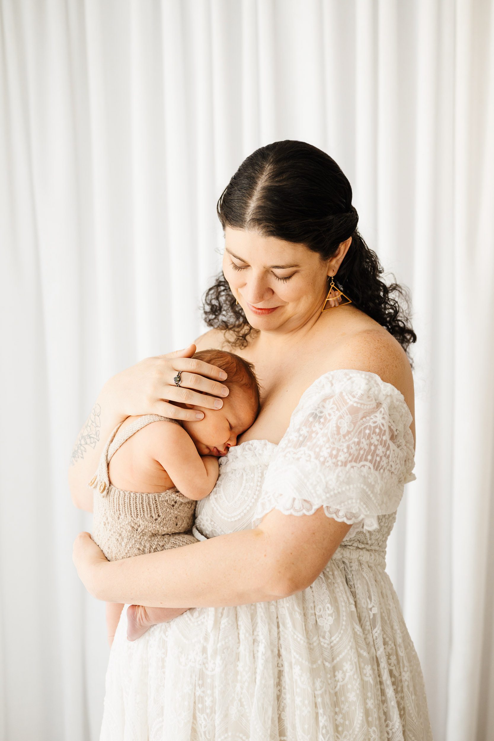 a new mom wearing a lacy white off the shoulder dress holding her baby boy snuggled up against her chest during a newborn photoshoot
