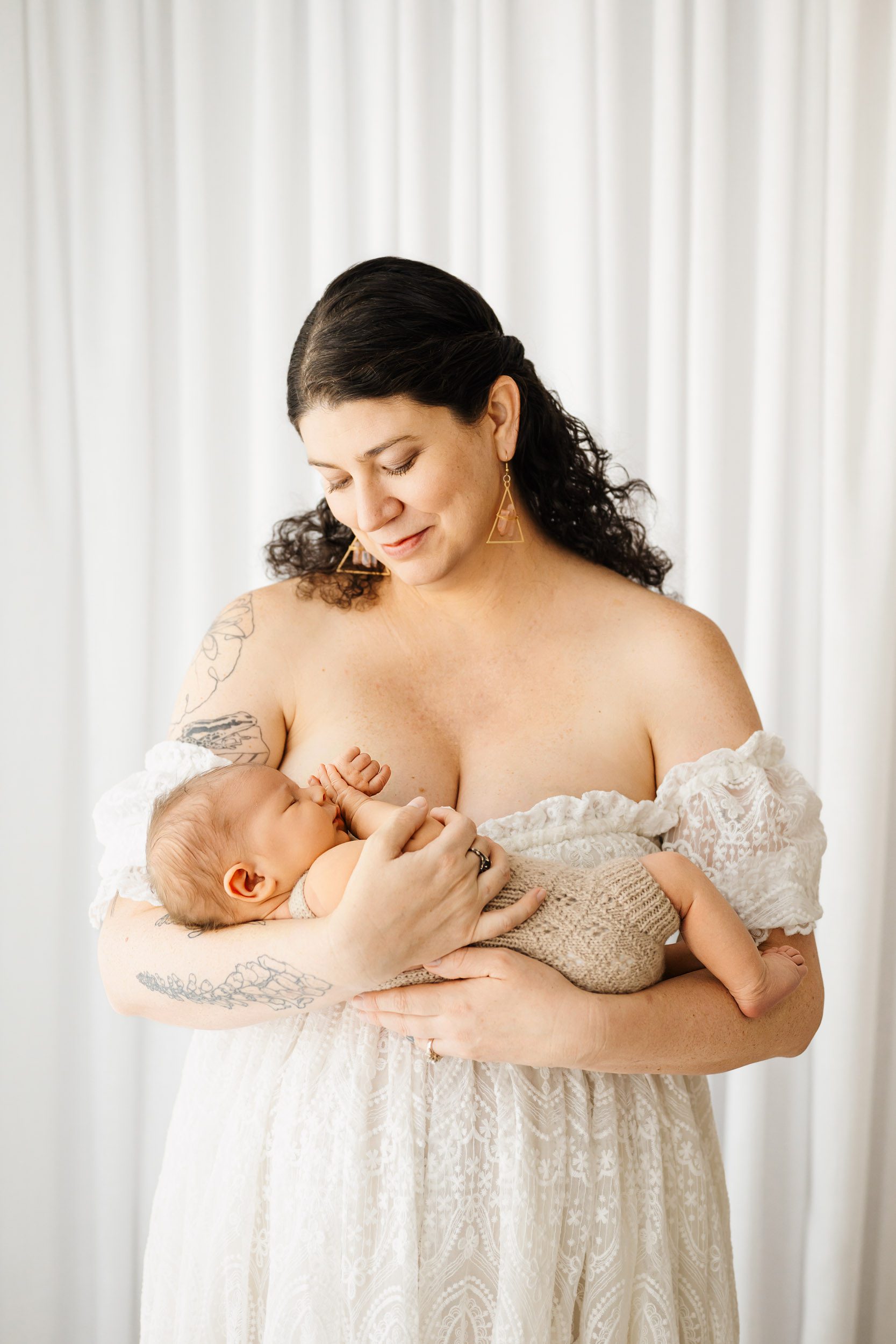 a new mom wearing a lacy white off the shoulder dress cradling her baby boy in her arms and smiling down at him during a newborn photoshoot