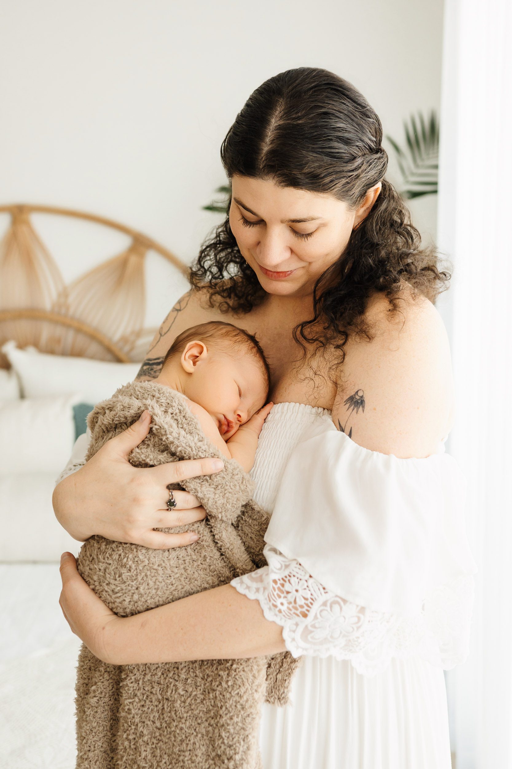 a new mom wearing a lacy white off the shoulder dress holding her baby boy snuggled up against her chest as he sleeps peacefully during a newborn photoshoot